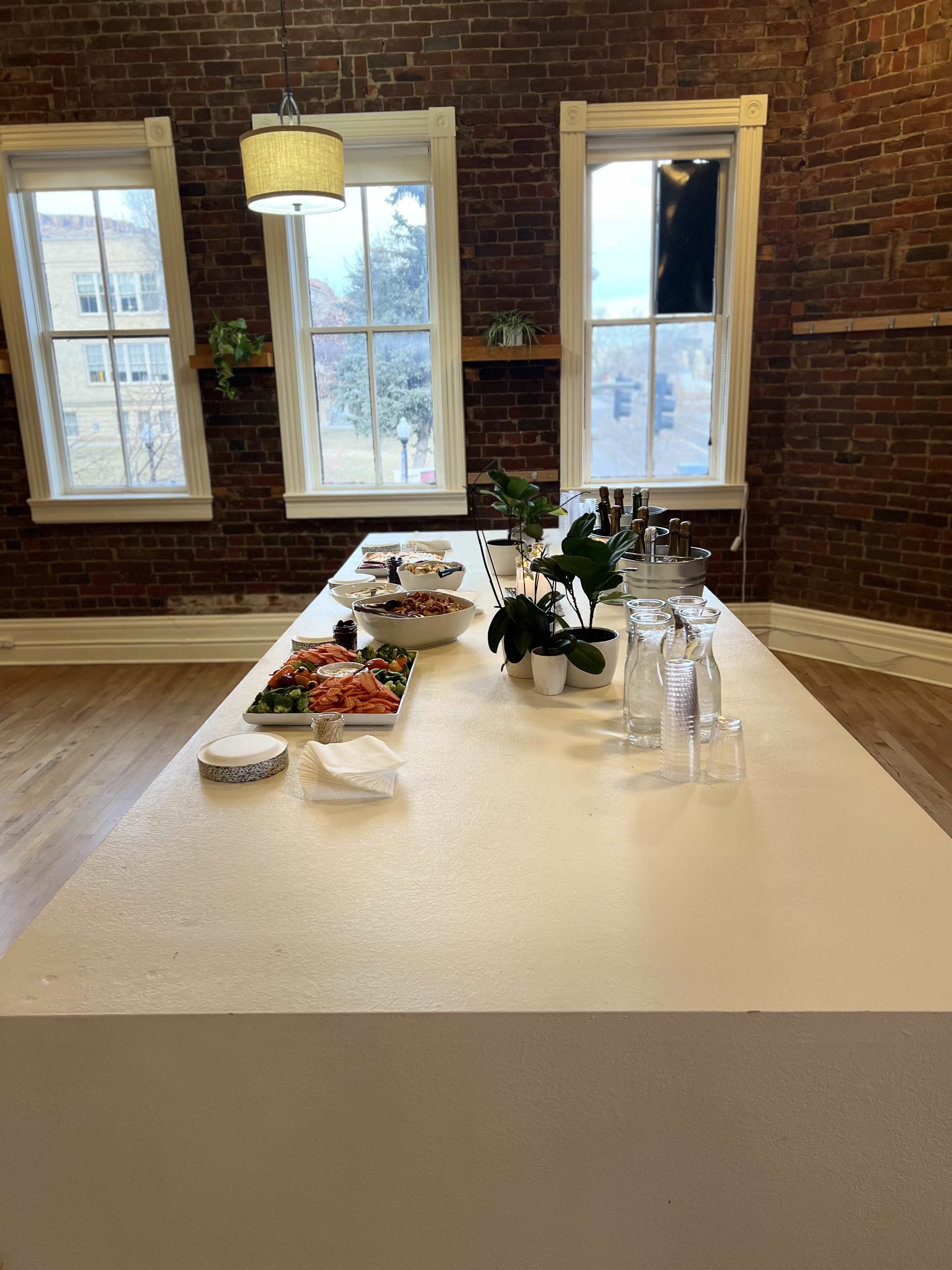 A white countertop displays a spread of food and drinks, surrounded by large windows framed by brick walls.
