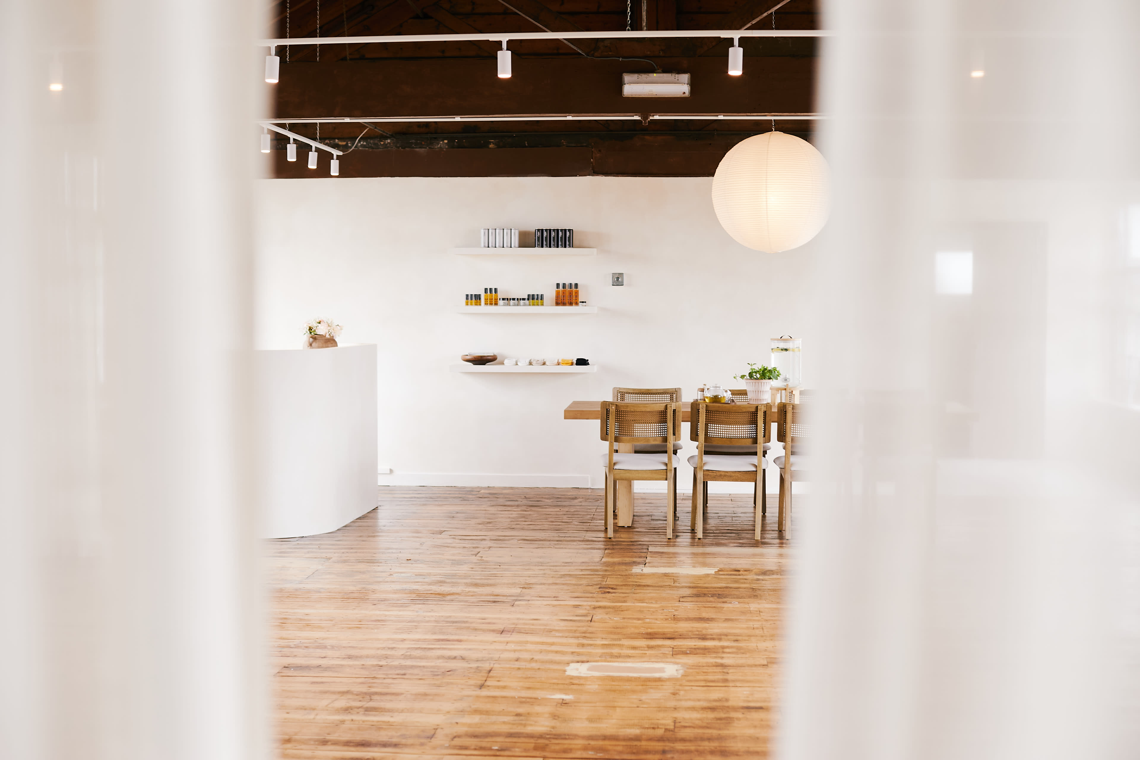 The image shows a serene interior space featuring a white reception desk, a wooden dining table with chairs, and shelves displaying various products, all illuminated by soft lighting.