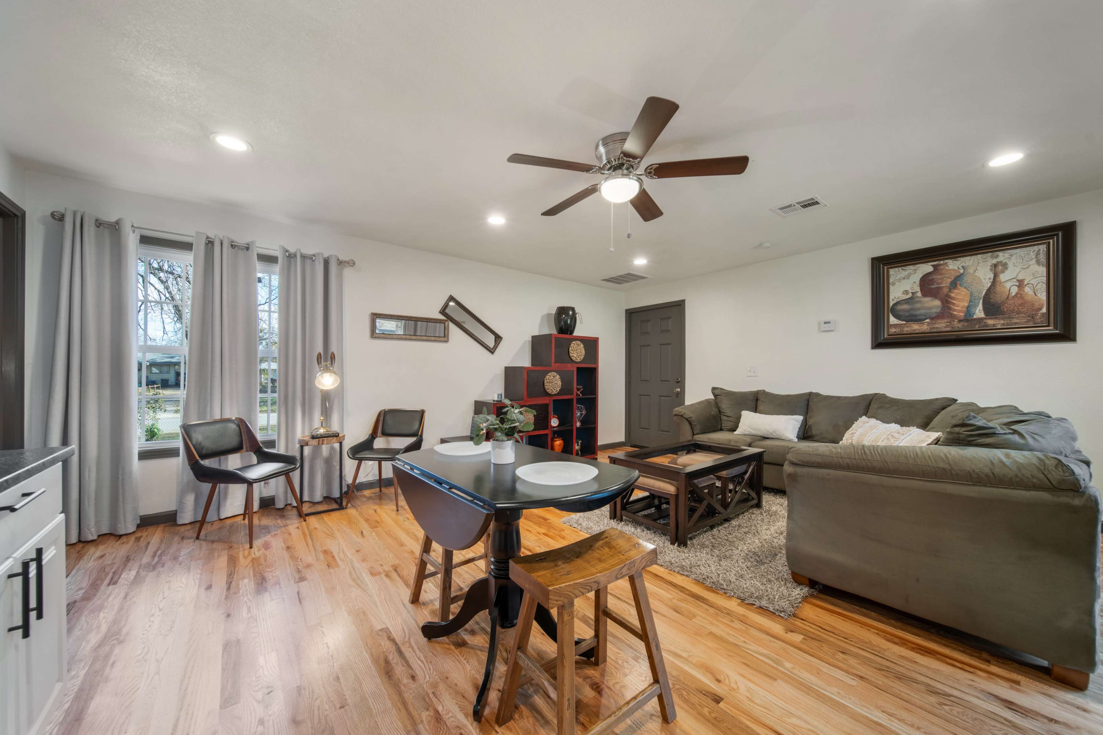 A living area features a sofa, a dining table with chairs, and decorative pieces, with natural light coming through a window.