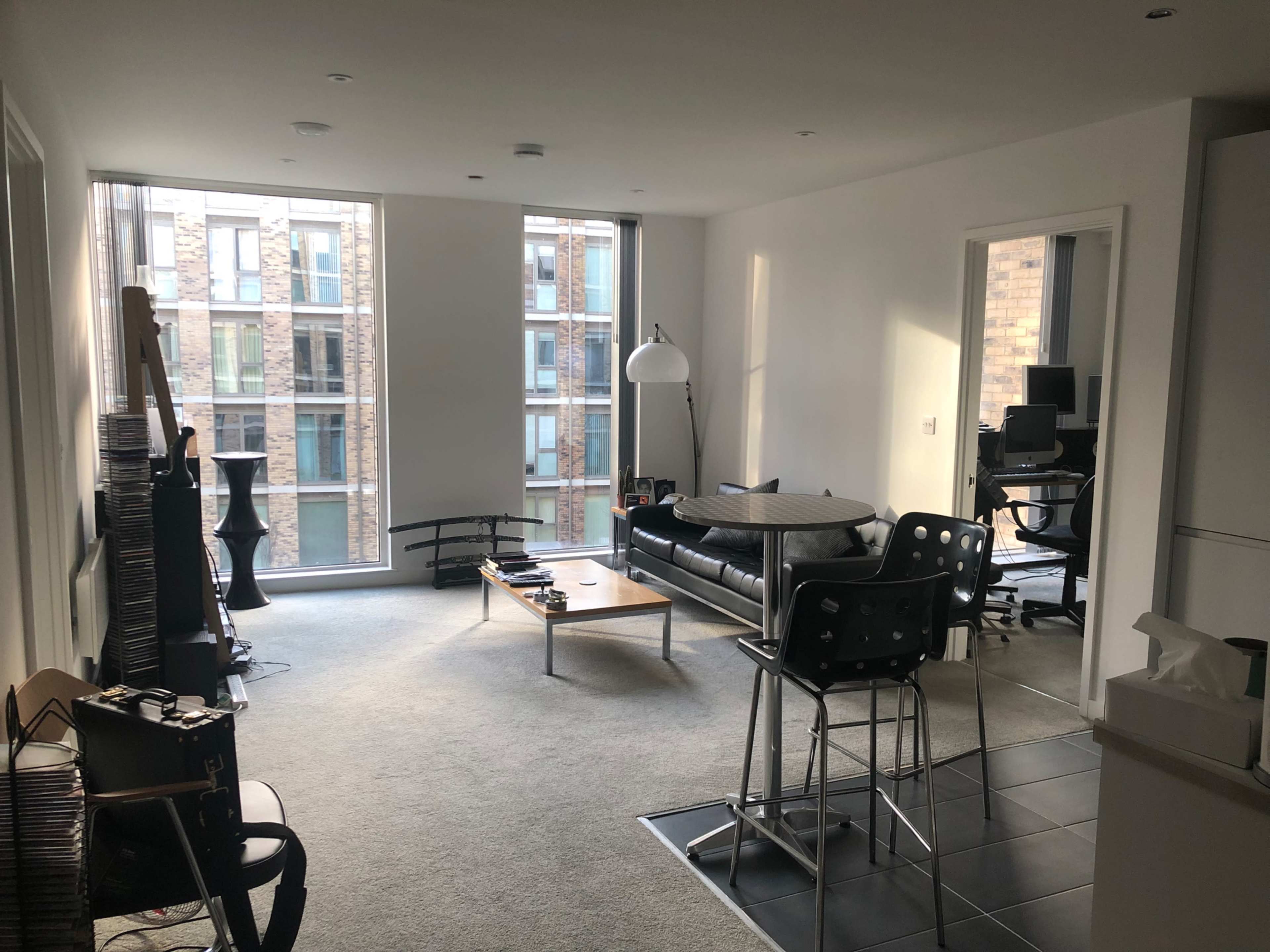 The image shows a spacious living room with large windows, a black sofa, a coffee table, and a workspace in the corner.