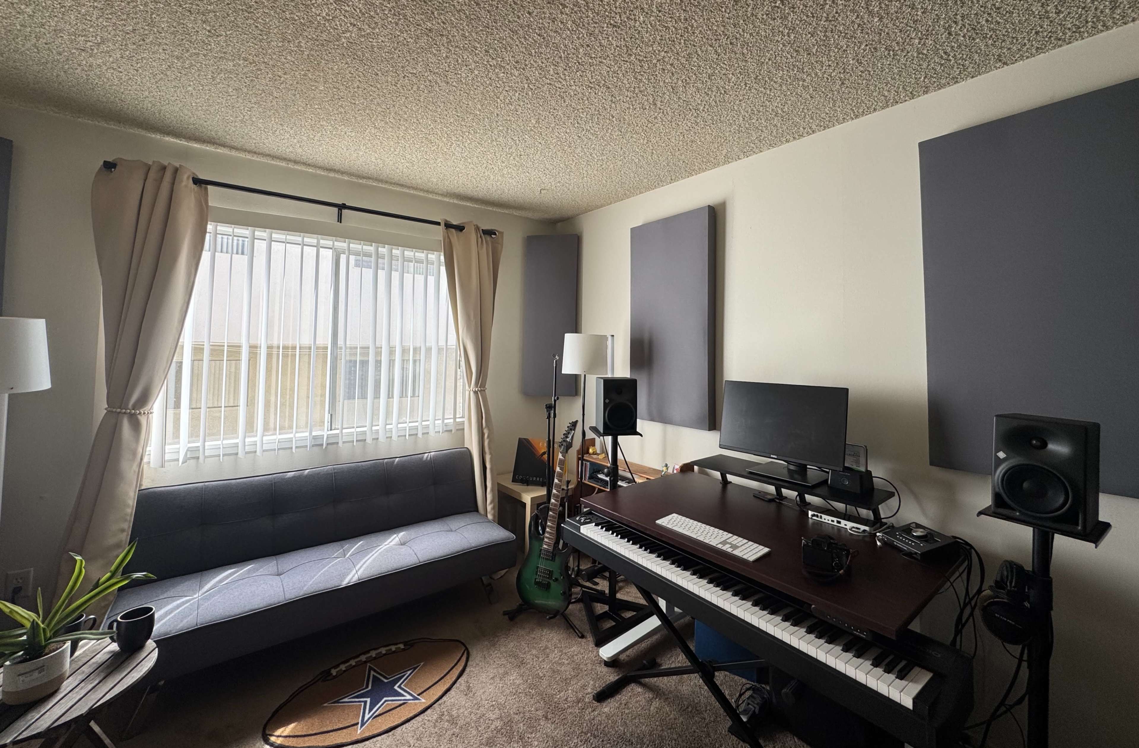 The image shows a small music studio with a keyboard, computer monitor, speakers, and a sofa, along with soundproofing panels on the walls and a window with curtains.