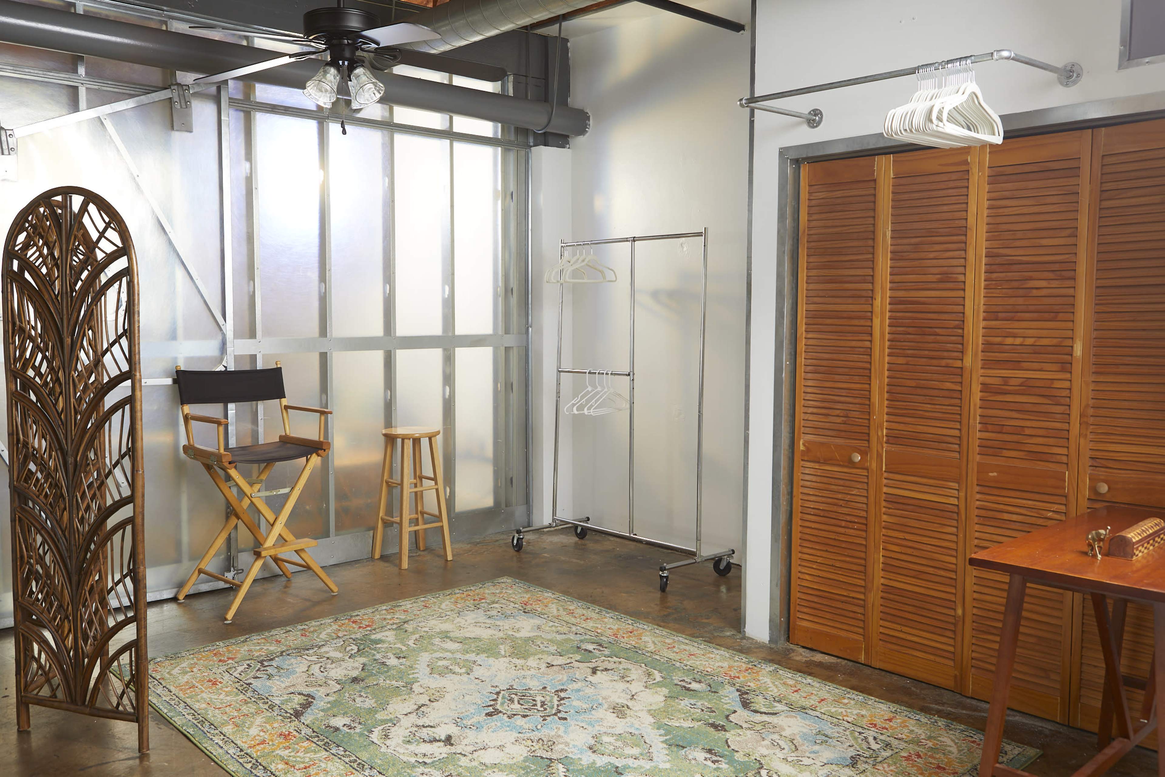 The image depicts a spacious room featuring a wooden folding screen, a director's chair, a stool, a clothing rack, and a wooden cabinet with louvered doors, all arranged on a decorative area rug.