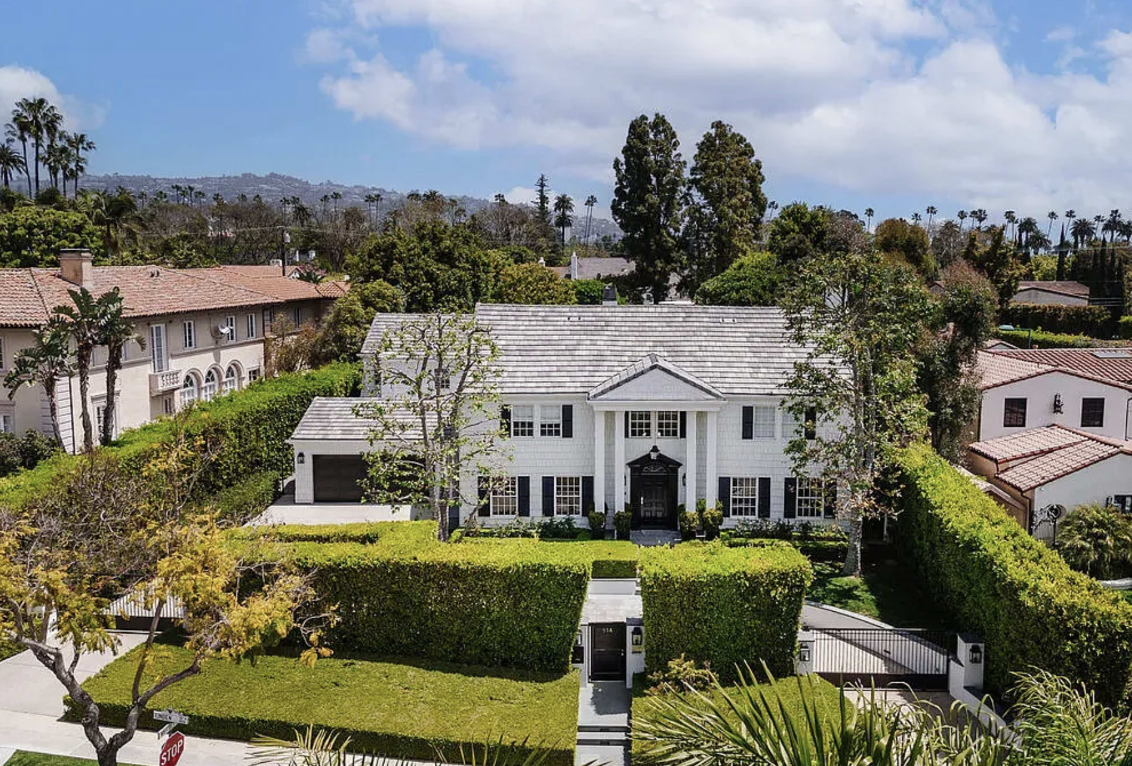 The image shows a large, two-story white house surrounded by well-manicured greenery and trees, with a clear blue sky in the background.