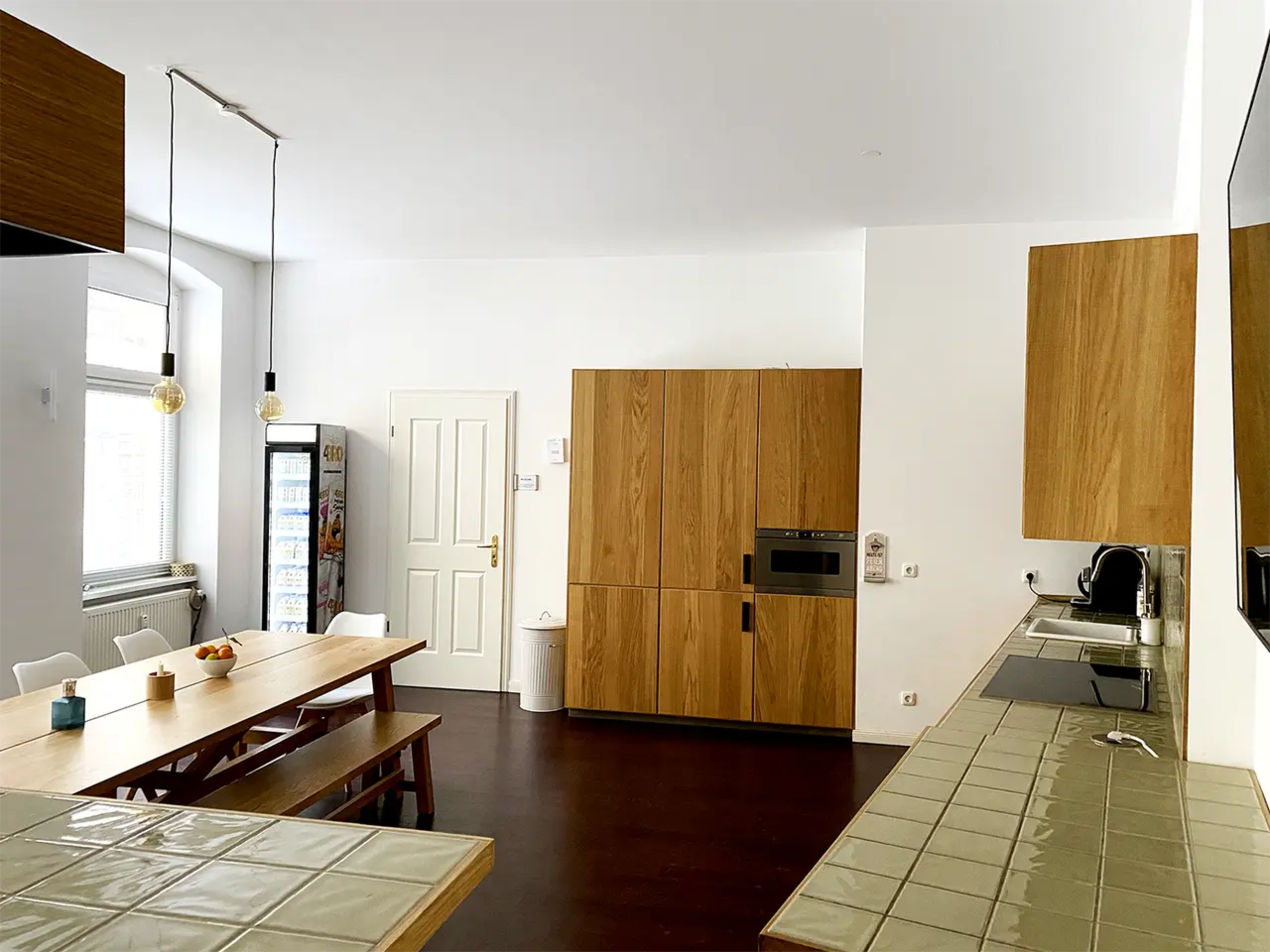 A spacious kitchen features wooden cabinetry, a long dining table, and modern appliances against a white wall.