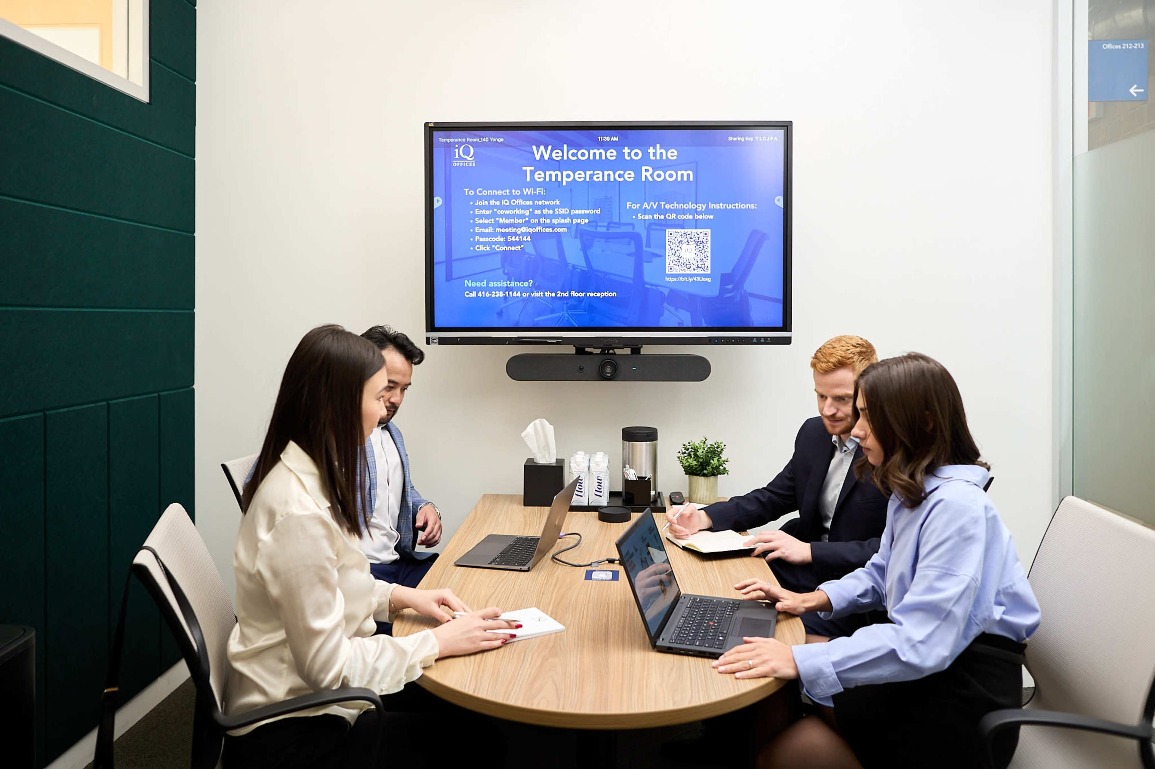 A group of four people is seated around a conference table in a meeting room, with a screen displaying a welcome message on the wall.
