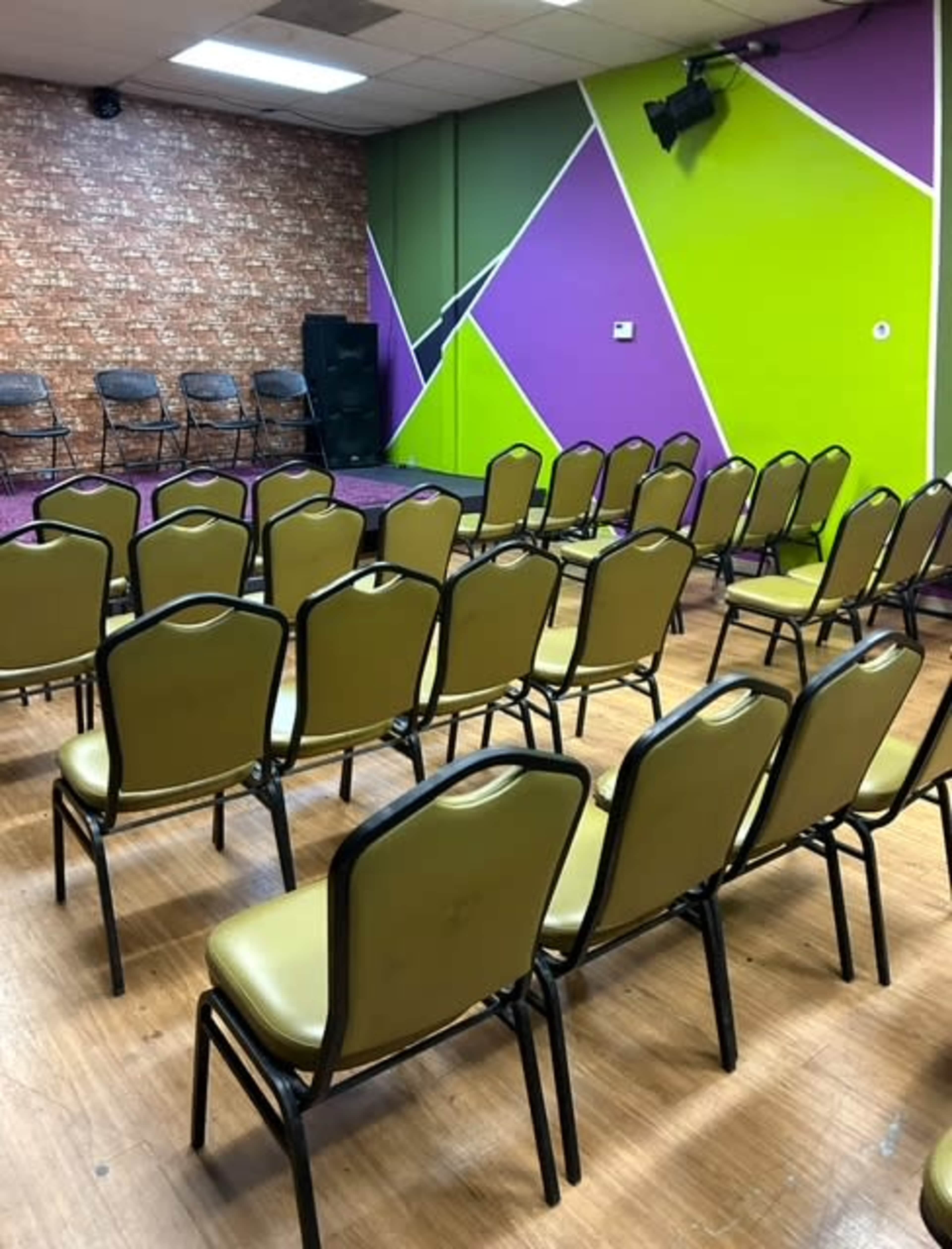The image shows a room with a stage and rows of empty green and black chairs arranged for an event, set against a colorful wall with geometric designs.