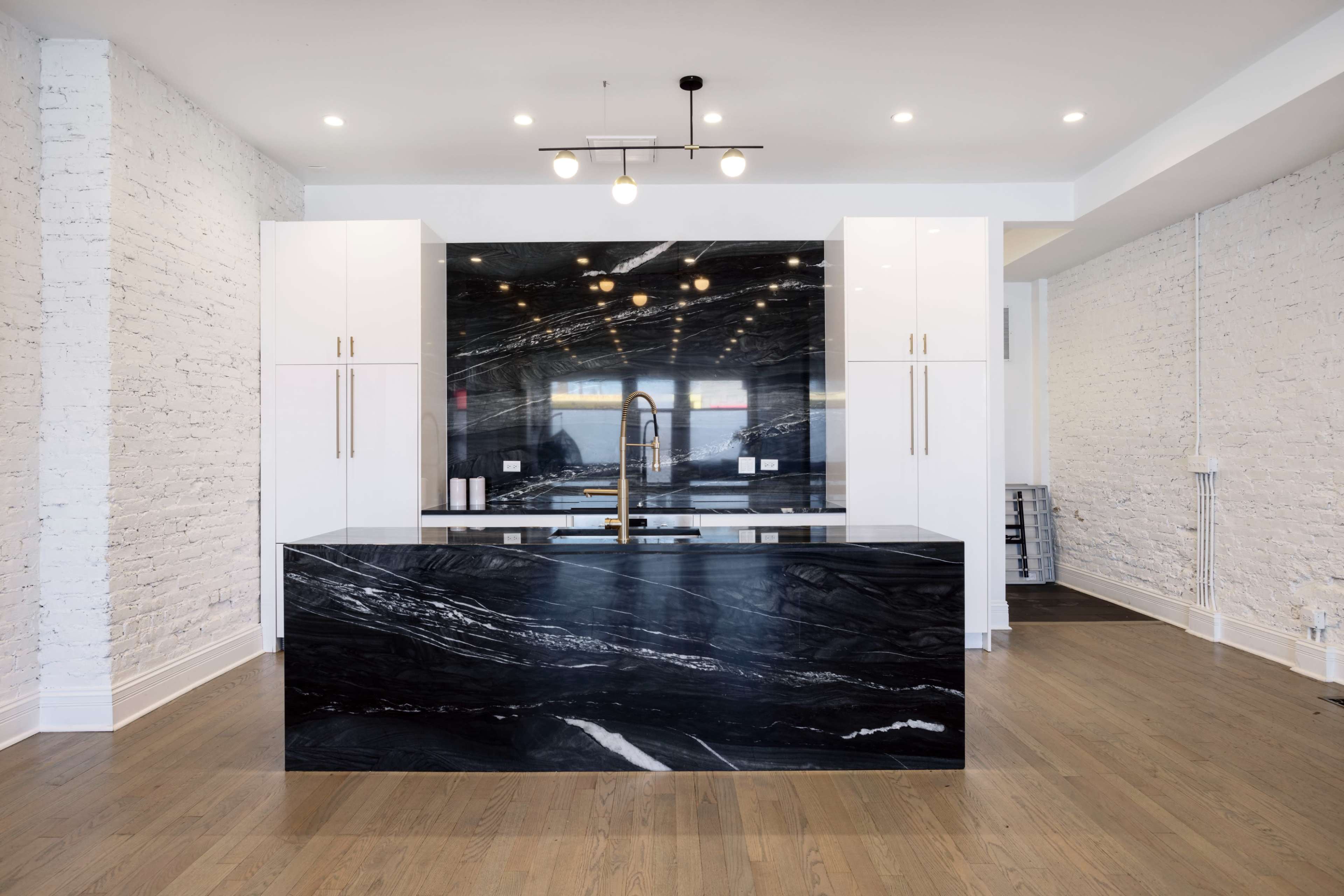 The image shows a modern kitchen with a large black marble island, white cabinets, and exposed white brick walls.