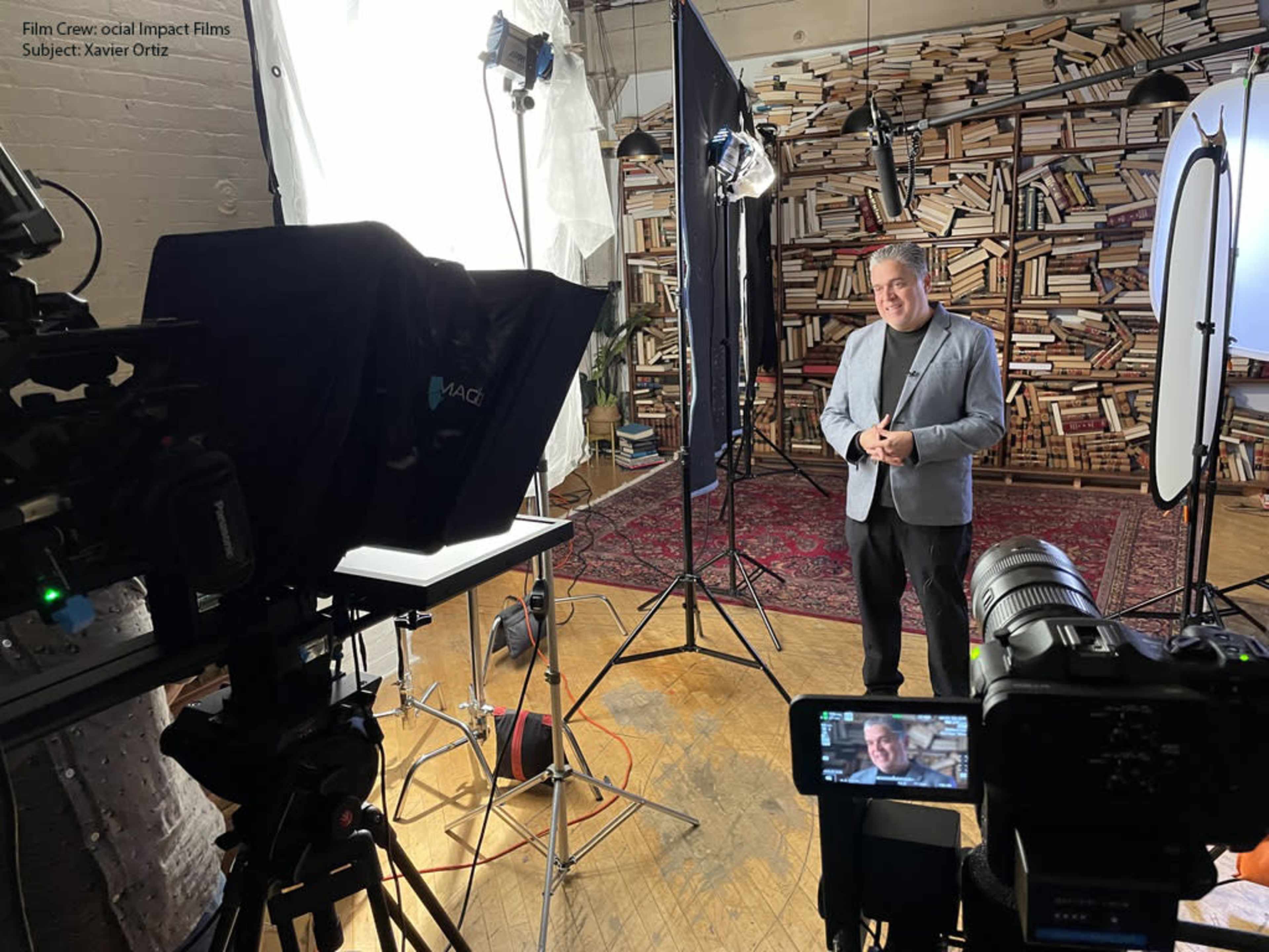 A man in a gray blazer stands in a studio setting with multiple lights and cameras in front of a wall lined with books.