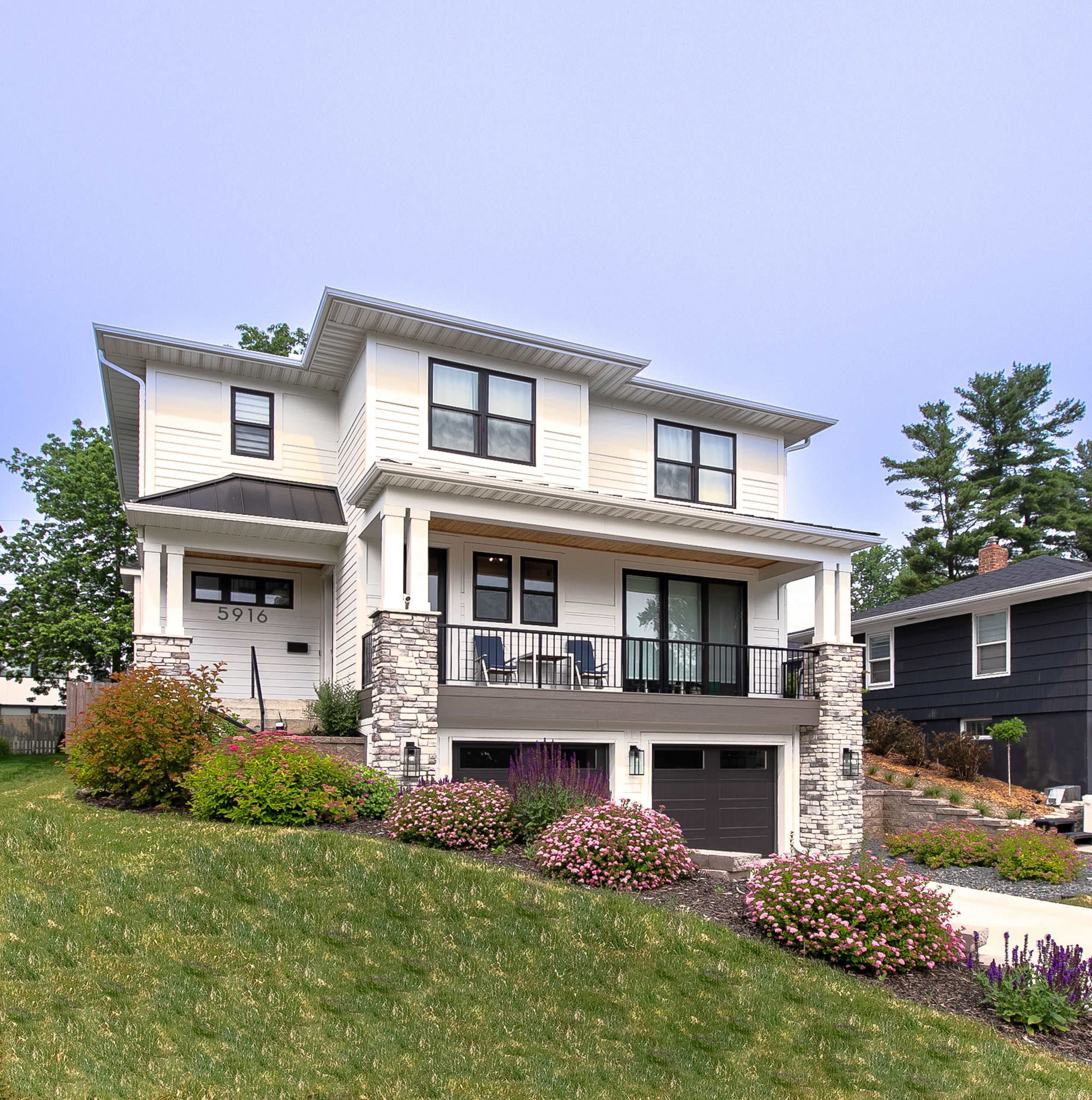 A two-story house with a front porch and stone accents sits on a landscaped hillside surrounded by flowering shrubs.