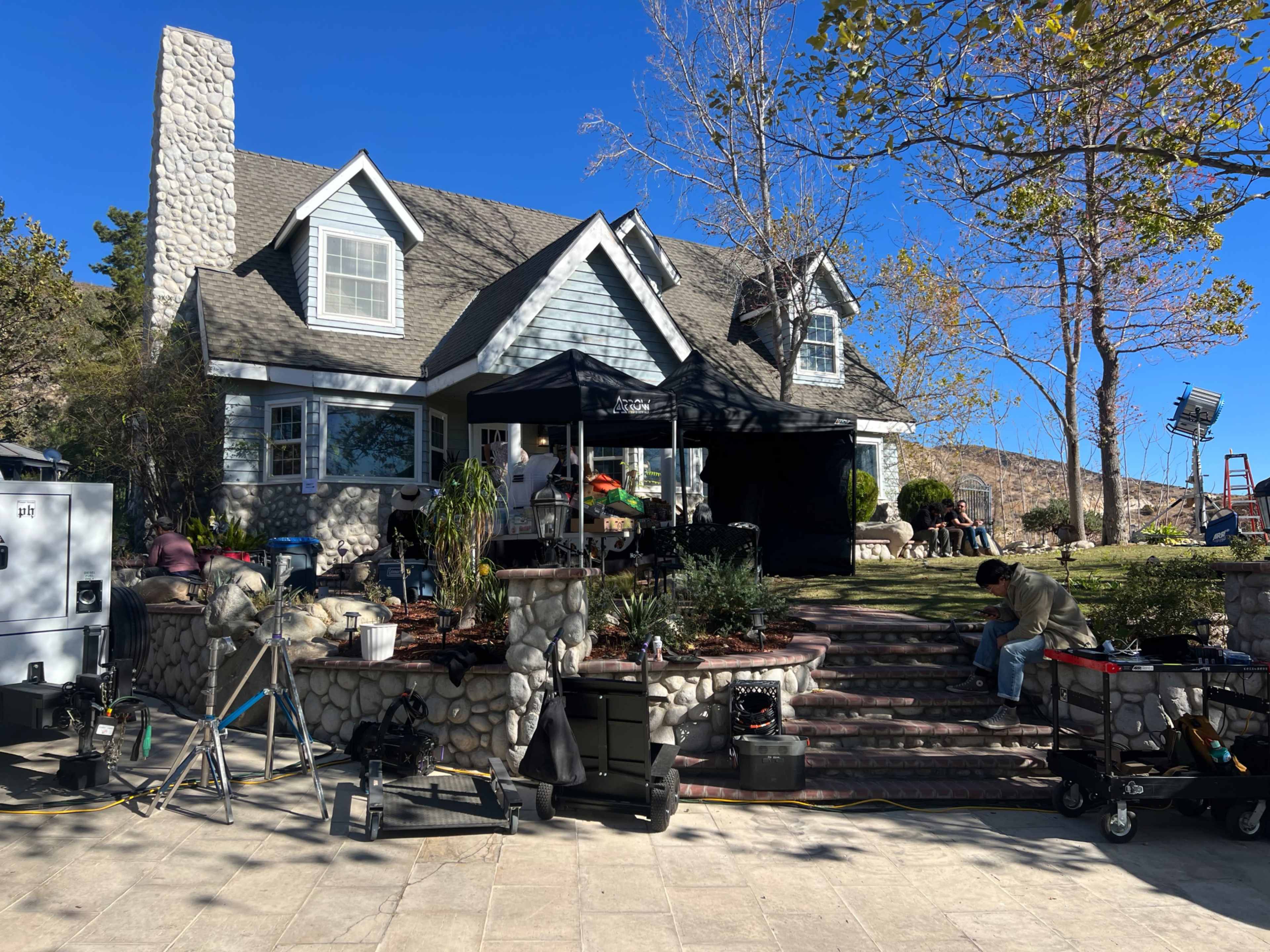 A film crew is set up in front of a house, with equipment and a canopy on a paved patio.