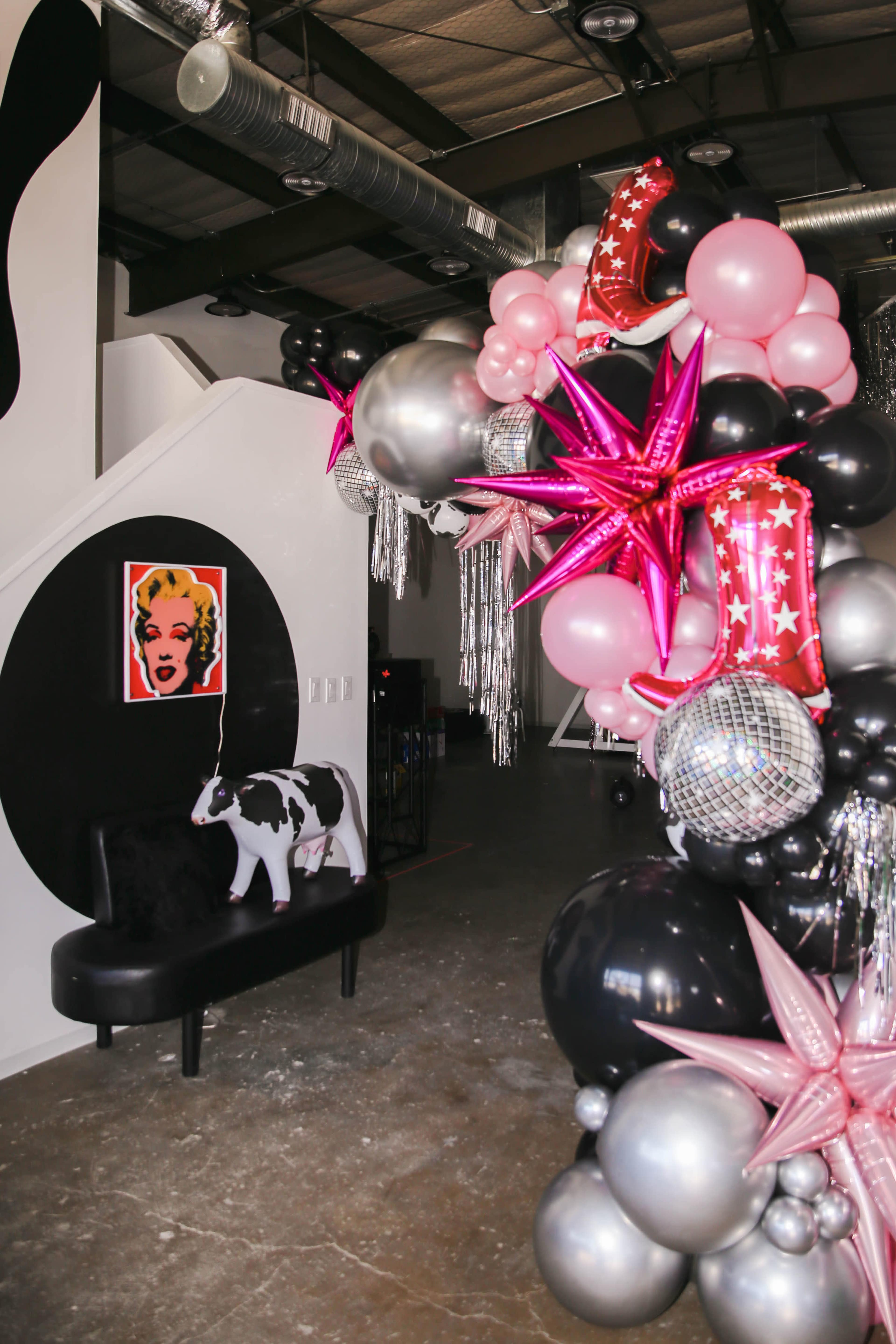 A colorful balloon arch featuring silver, pink, black, and star-shaped balloons leads to a seating area with a cow statue and a Marilyn Monroe poster on the wall.
