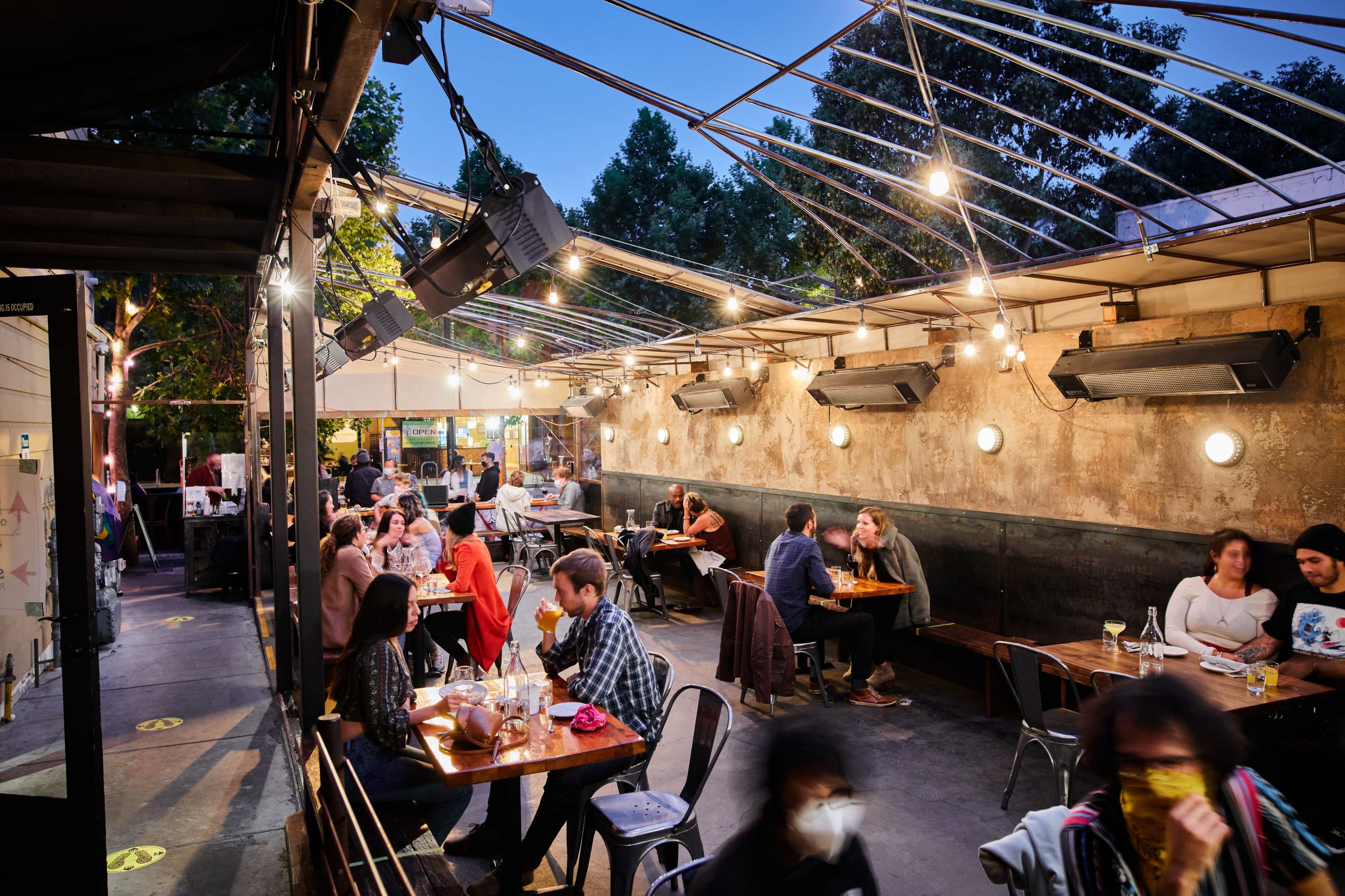 The image shows a lively outdoor dining area with string lights overhead, where groups of people are seated at tables enjoying their meals.
