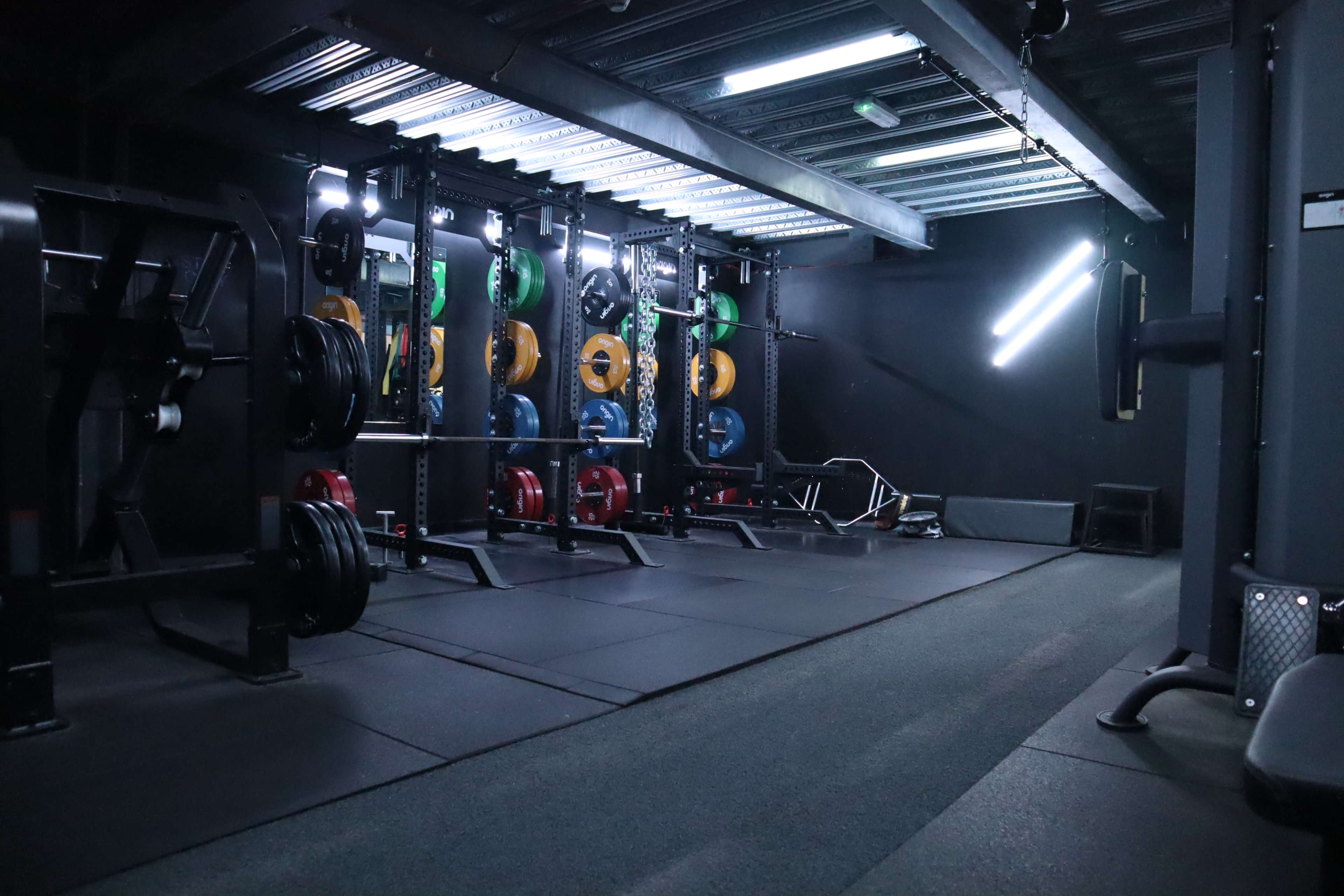 A dimly lit gym features multiple weightlifting stations equipped with colored weight plates and exercise benches.