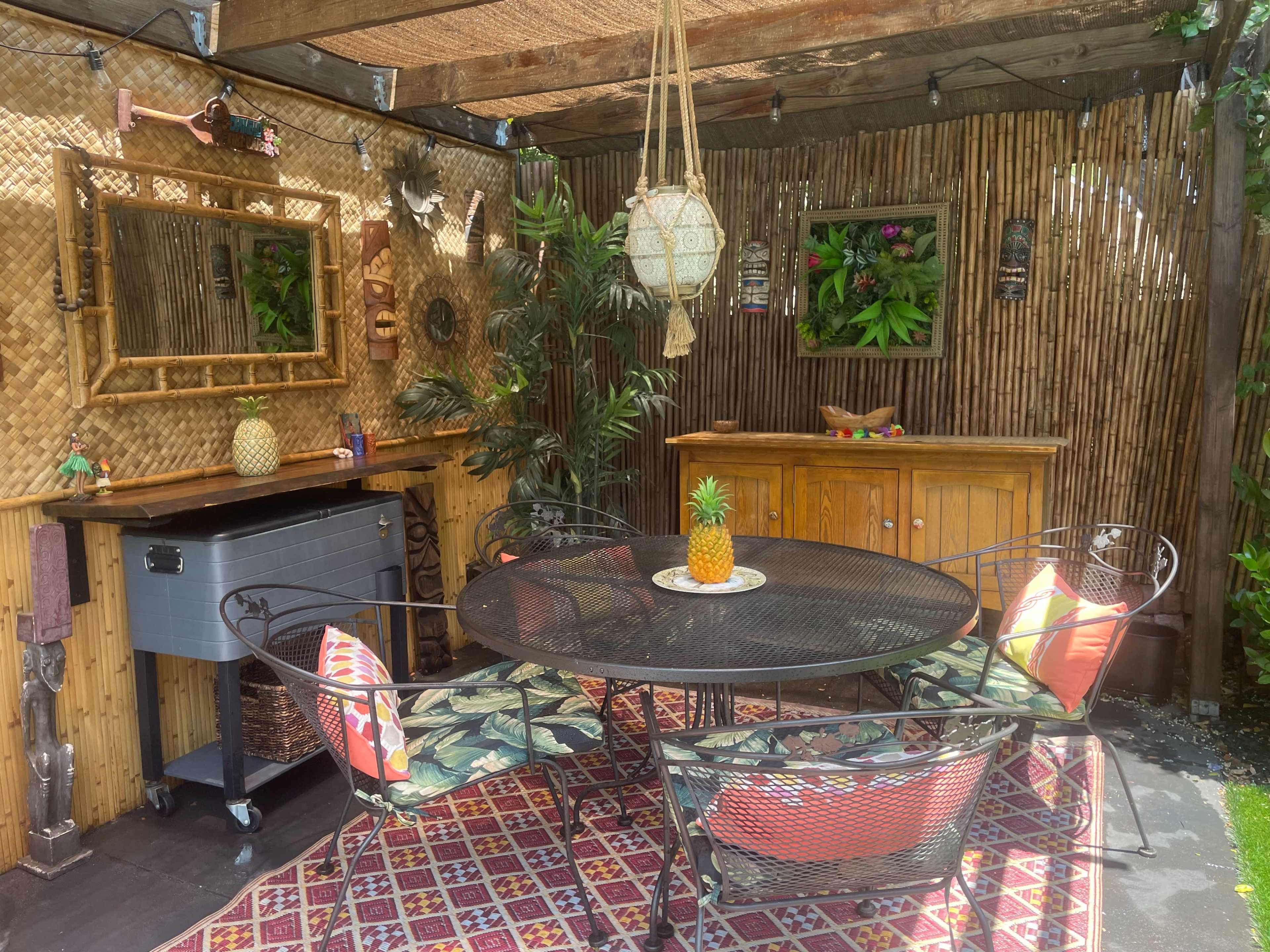 The image shows a shaded patio area furnished with a circular metal table surrounded by four chairs, a bamboo-lined bar, decorative tropical plants, and a patterned carpet.