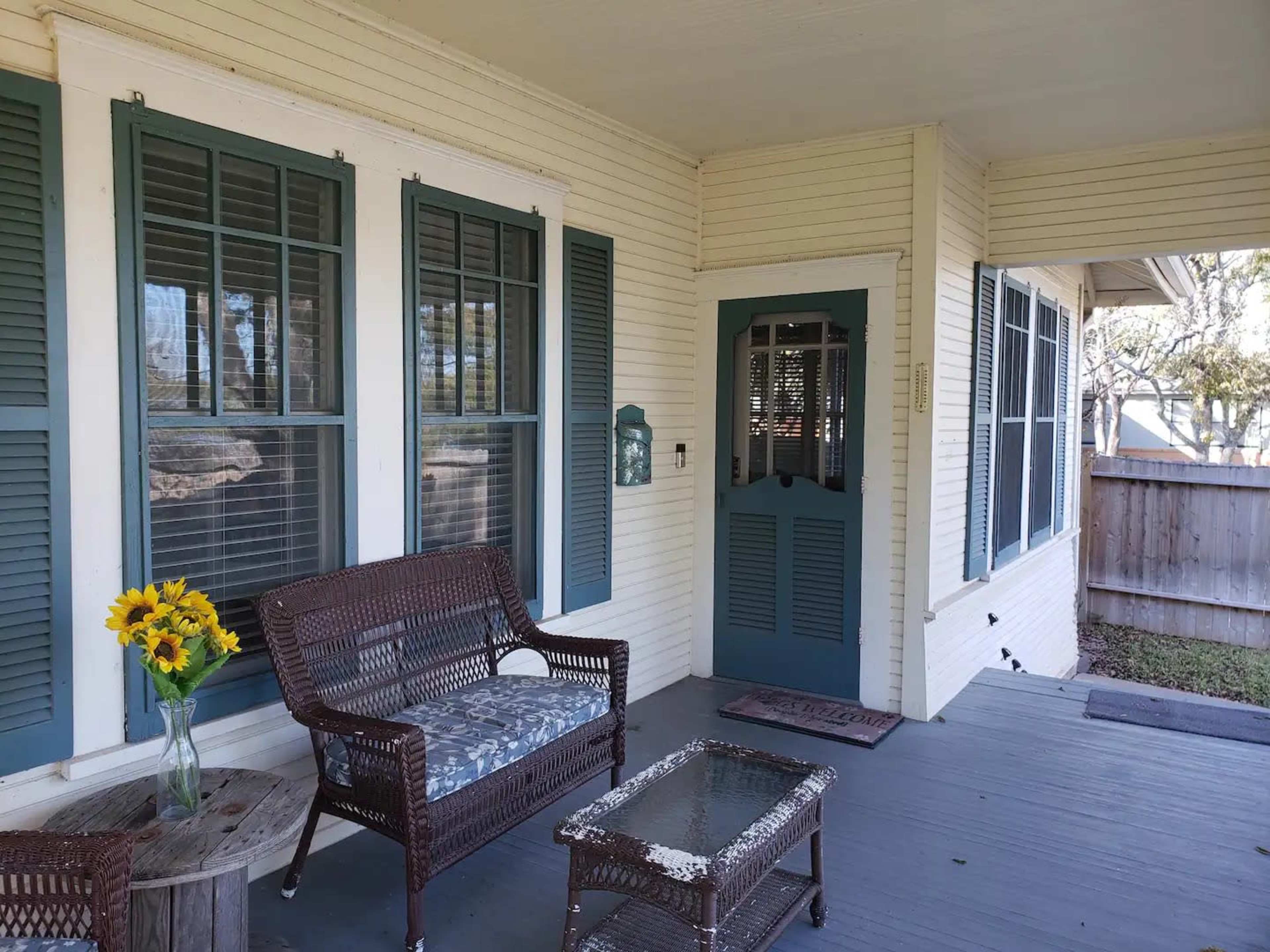 The image shows a porch with a wicker seating area, a small table, and a bright bouquet of sunflowers on it, alongside a door with a decorative frame.