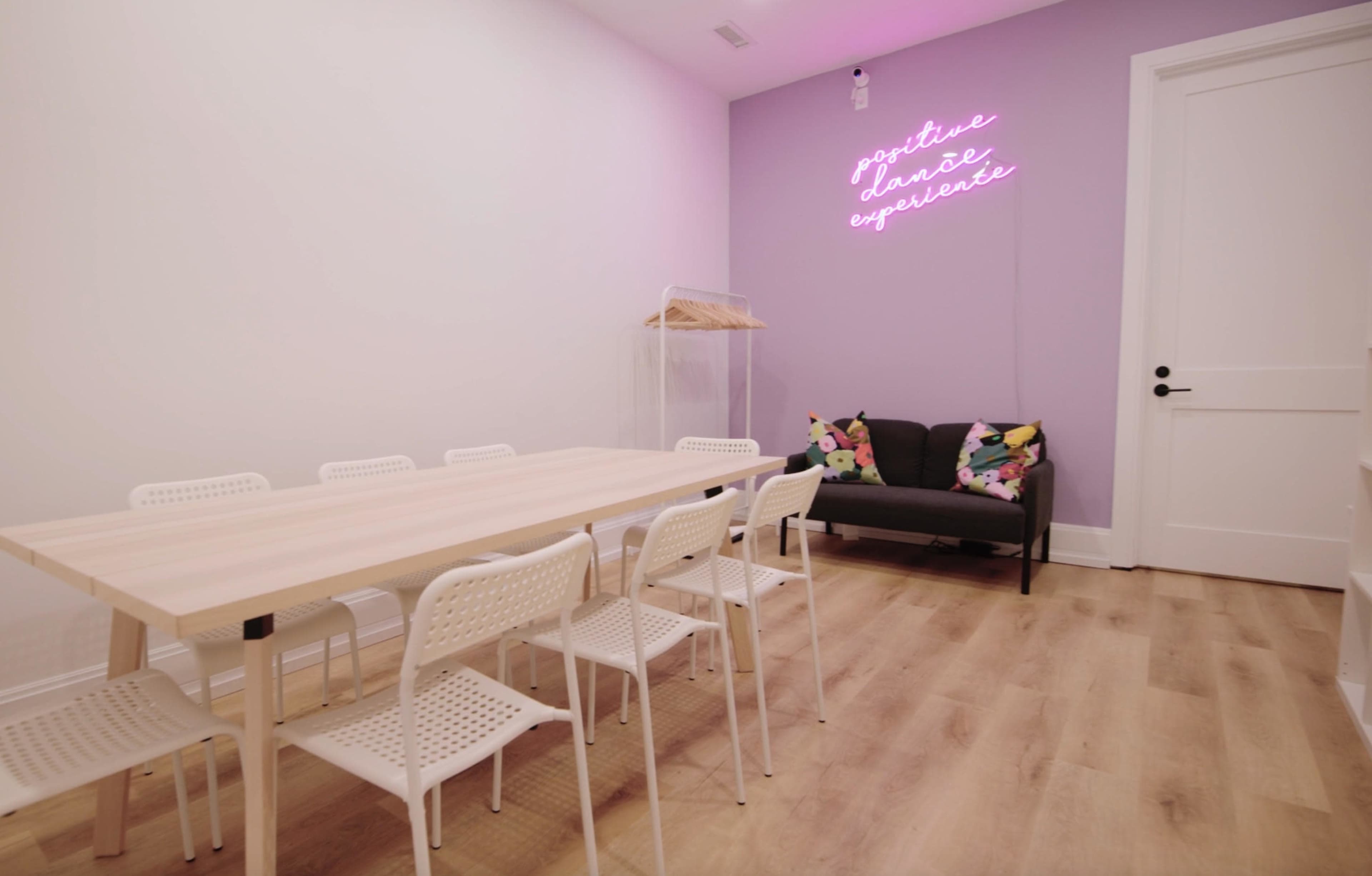 A minimalist meeting room features a long wooden table surrounded by white chairs, alongside a black sofa adorned with colorful pillows, against a pastel purple wall with a neon sign.