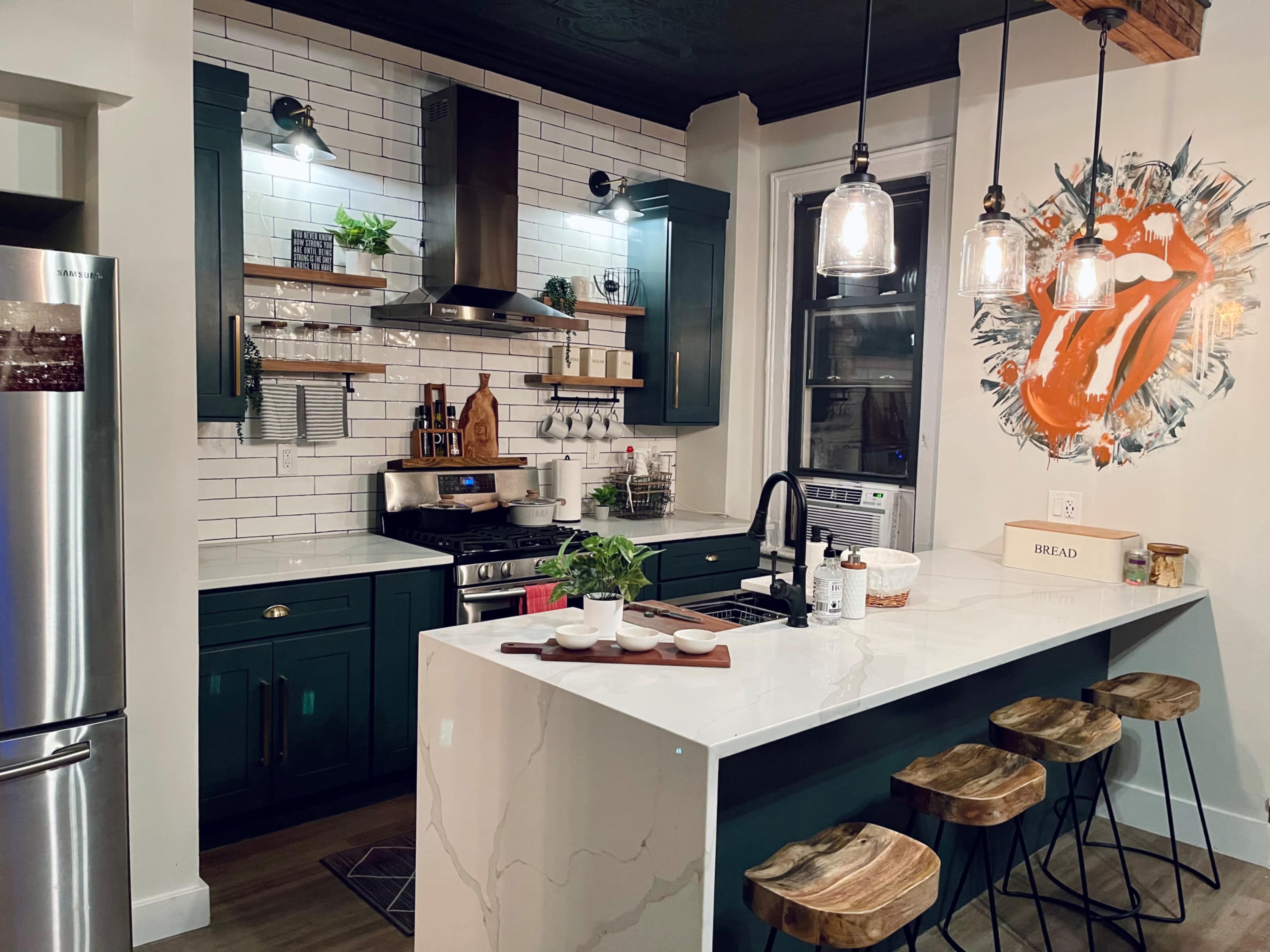 A modern kitchen featuring a central island with wooden stools, dark green cabinetry, white subway tile, and a bold orange art piece on the wall.