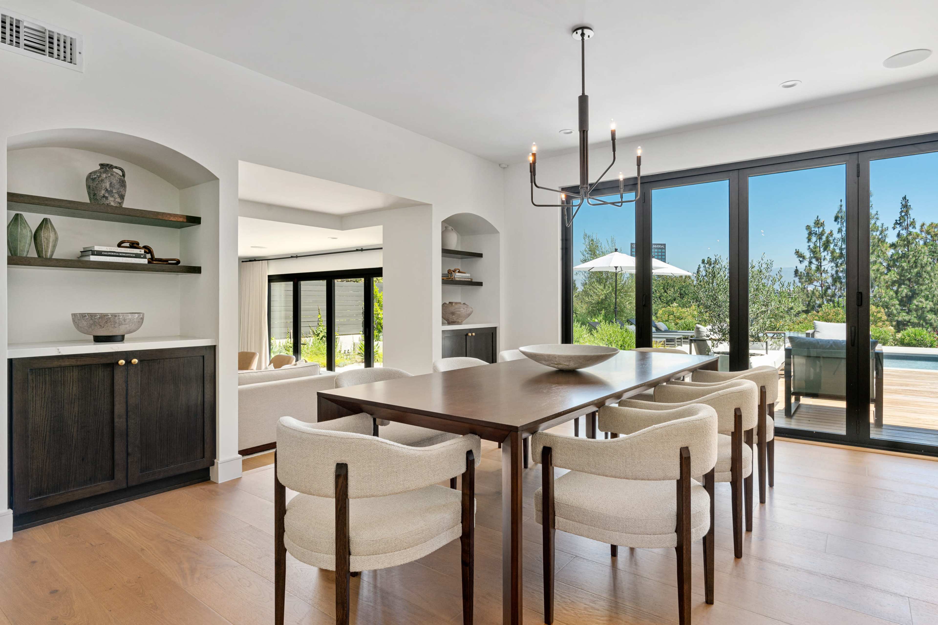 A dining area features a wooden table surrounded by cushioned chairs, with large glass doors leading to an outdoor space.