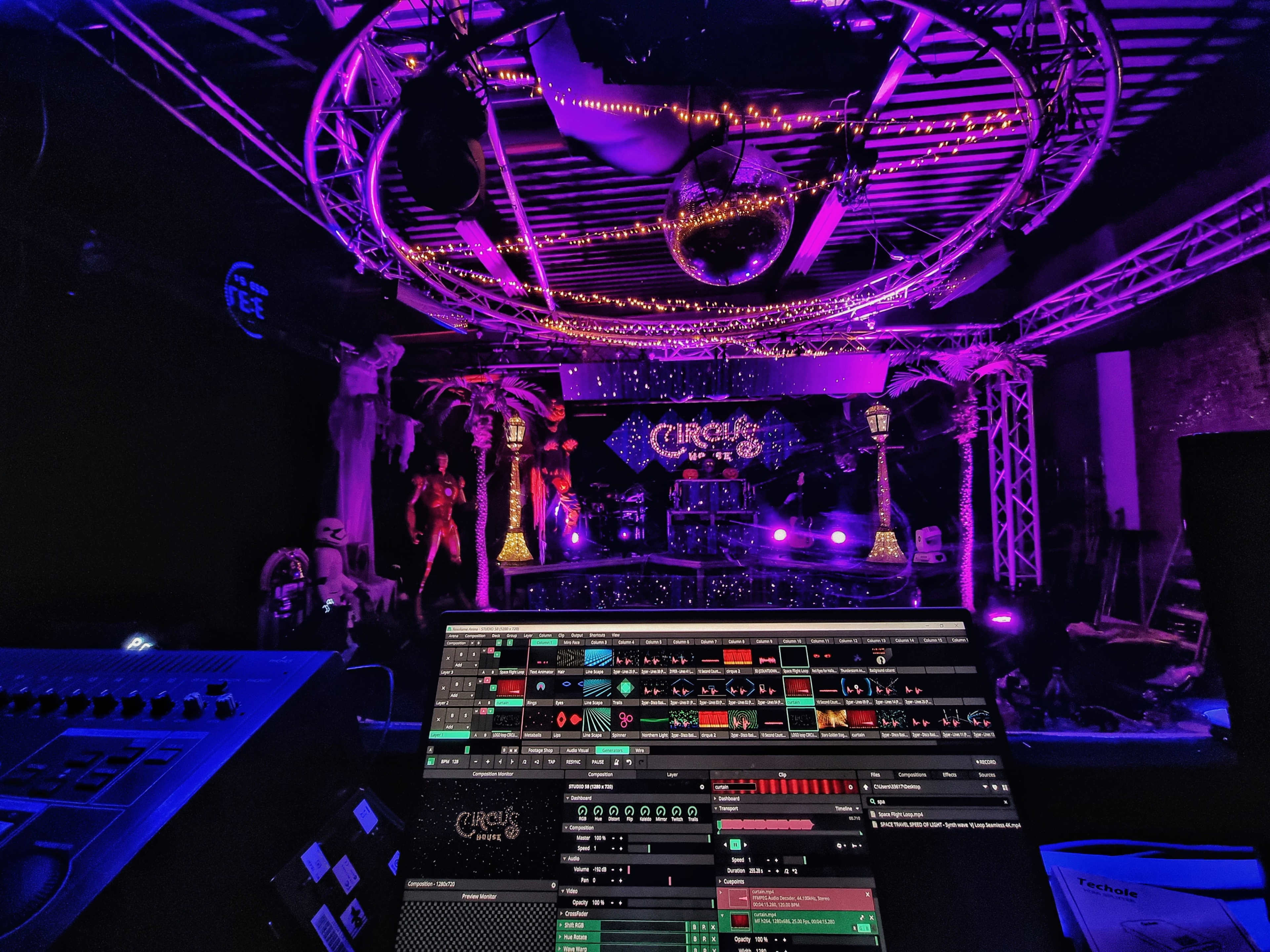 A stage set up for a circus-themed event, featuring decorative elements like palm trees, a disco ball, and vibrant purple lighting, with a sound mixing console in the foreground.