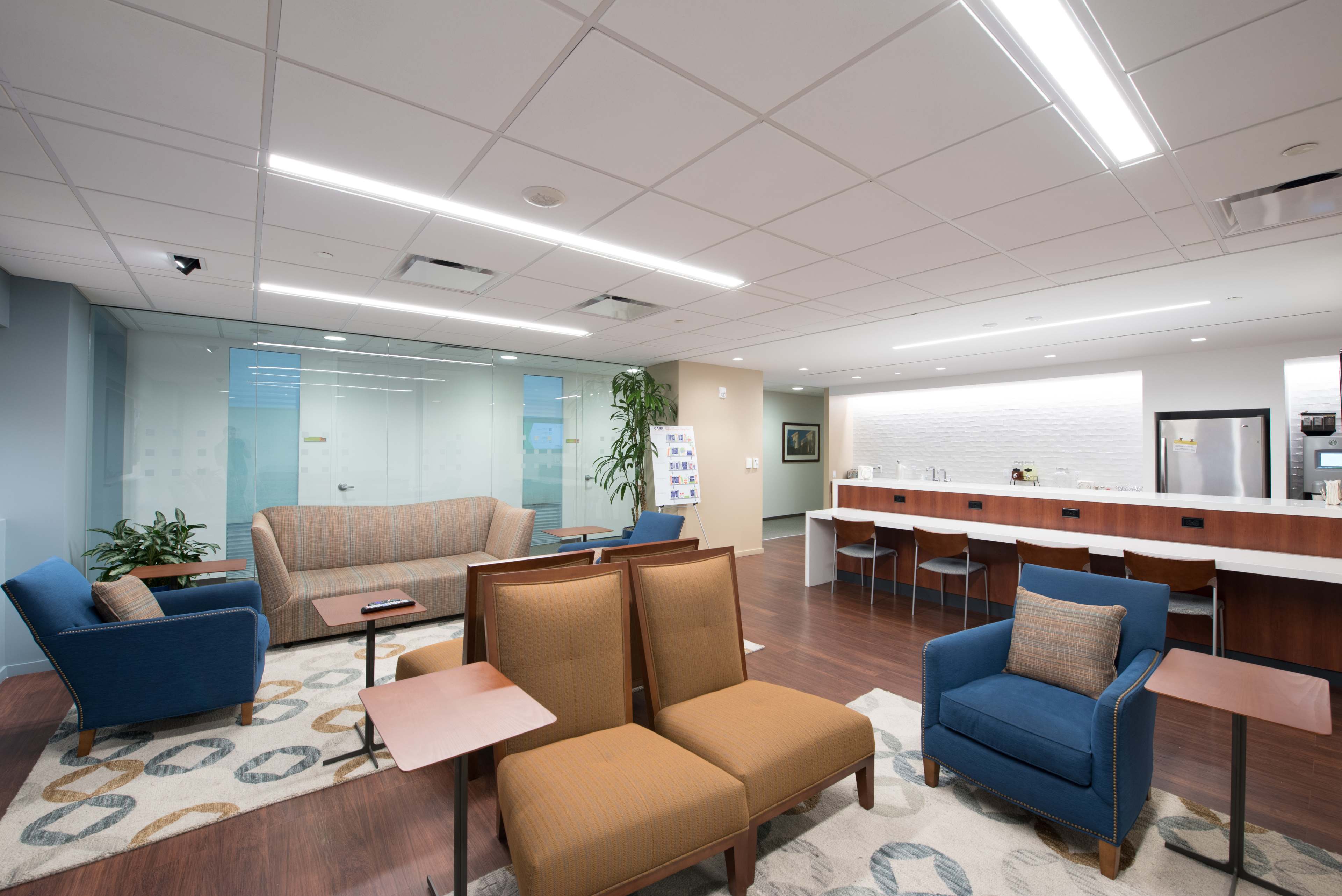 A modern lounge area with seating arrangements, including sofas and chairs, along with a kitchenette in a well-lit space.