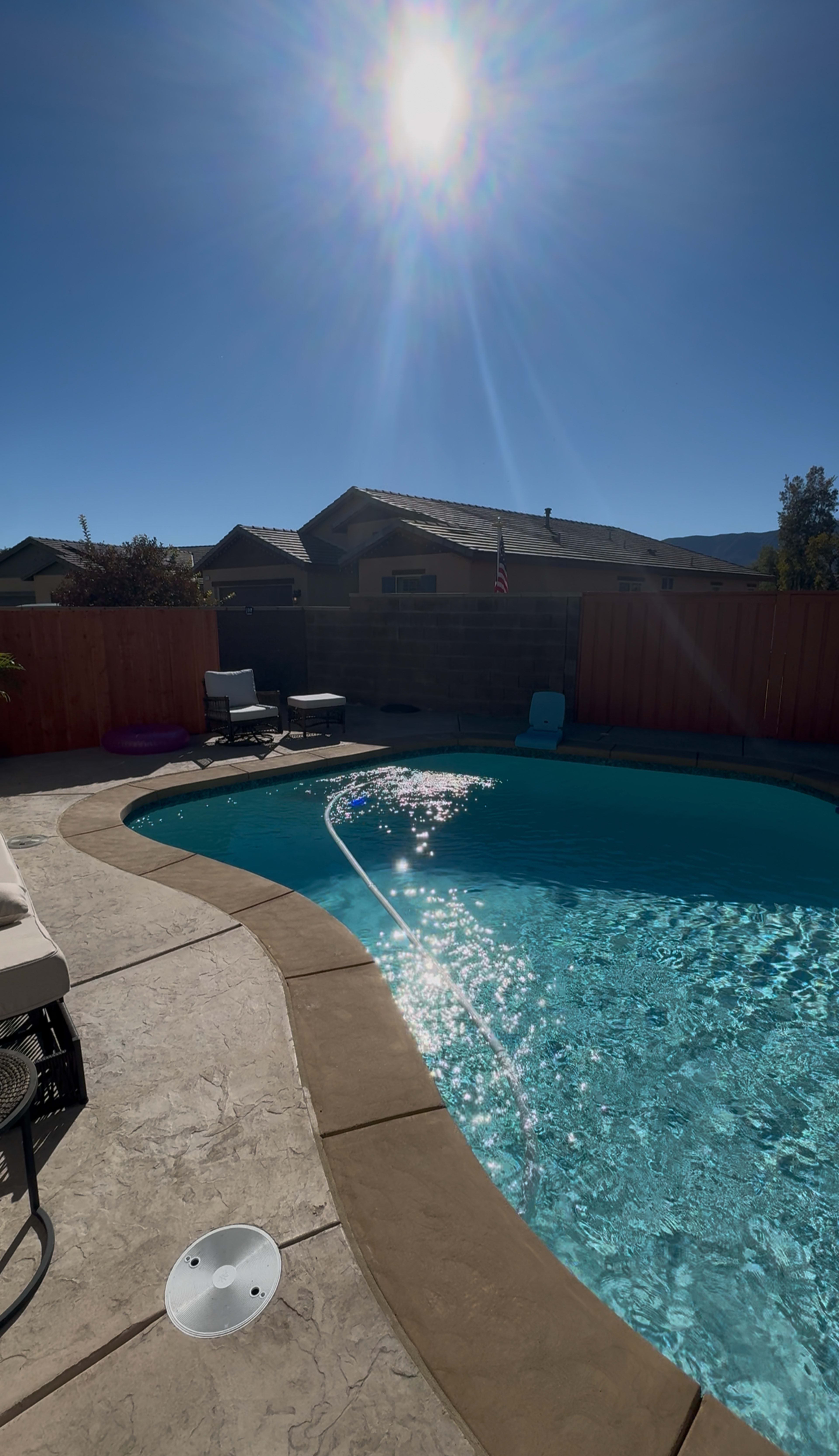 The image shows a clear pool surrounded by concrete and a wooden fence, with a house in the background and bright sunlight reflecting off the water.
