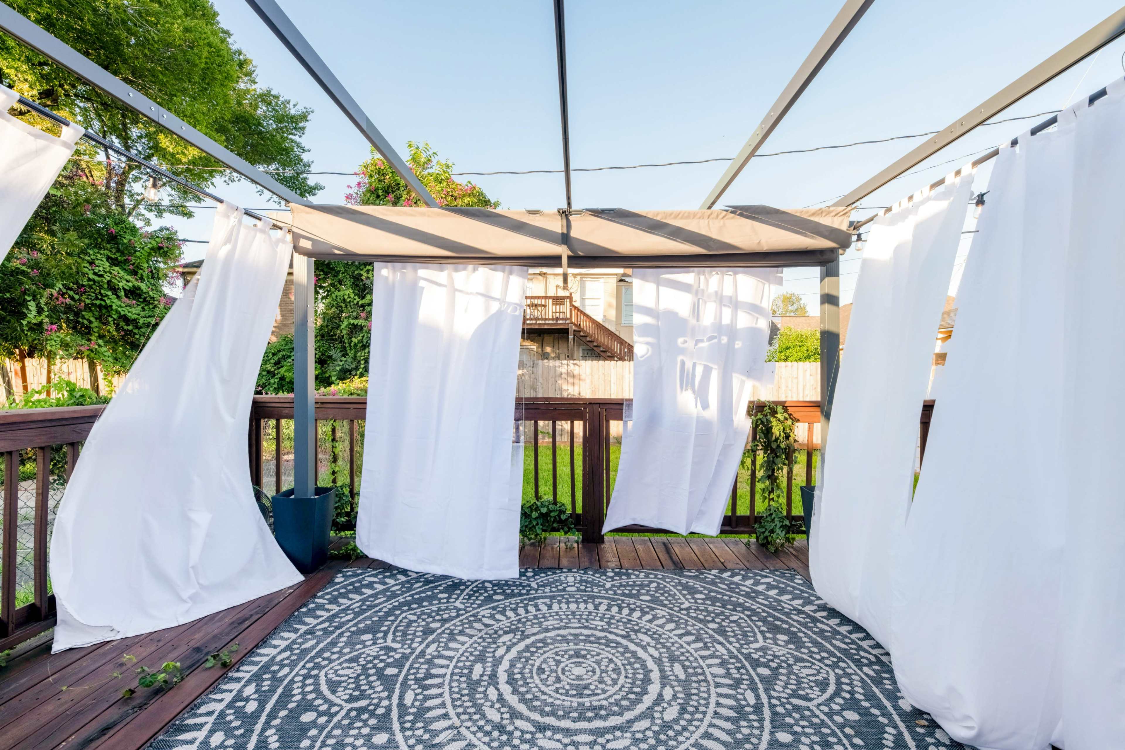 The image shows a wooden deck covered by a pergola with white curtains and a circular patterned rug on the floor.