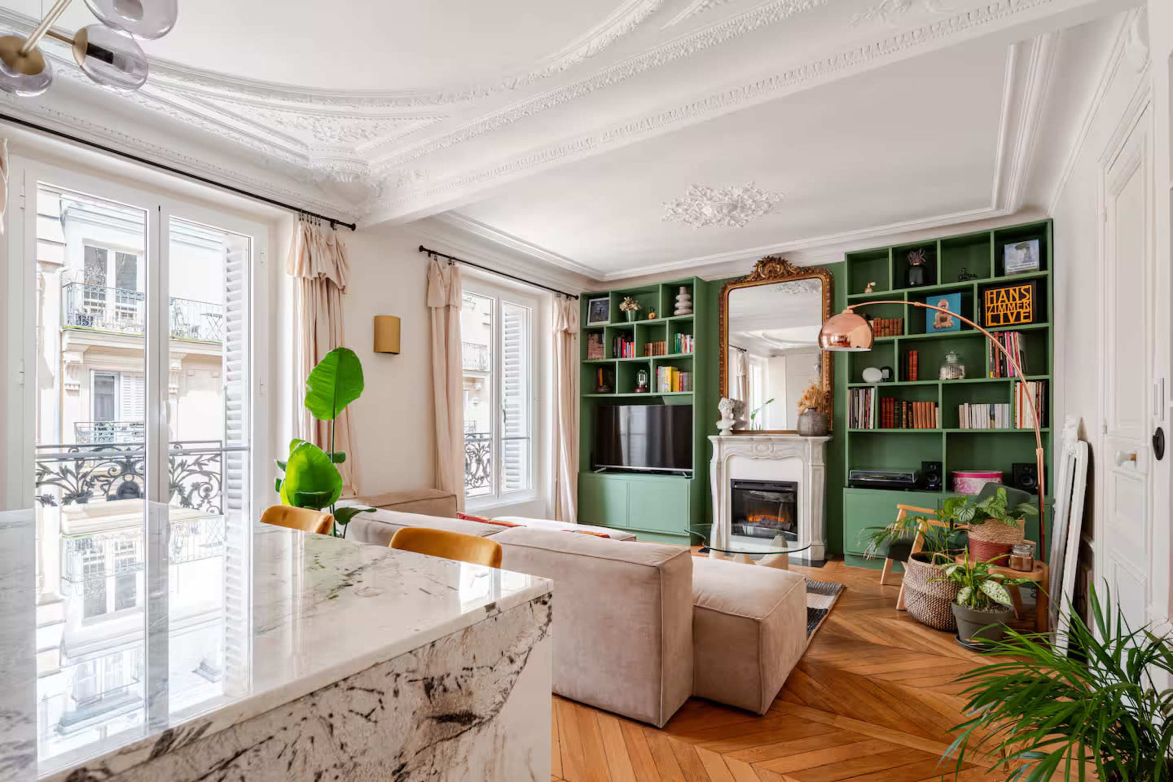 The image features a stylish living room with a beige sofa, a green bookshelf filled with books, and a large window that allows natural light to illuminate the space.