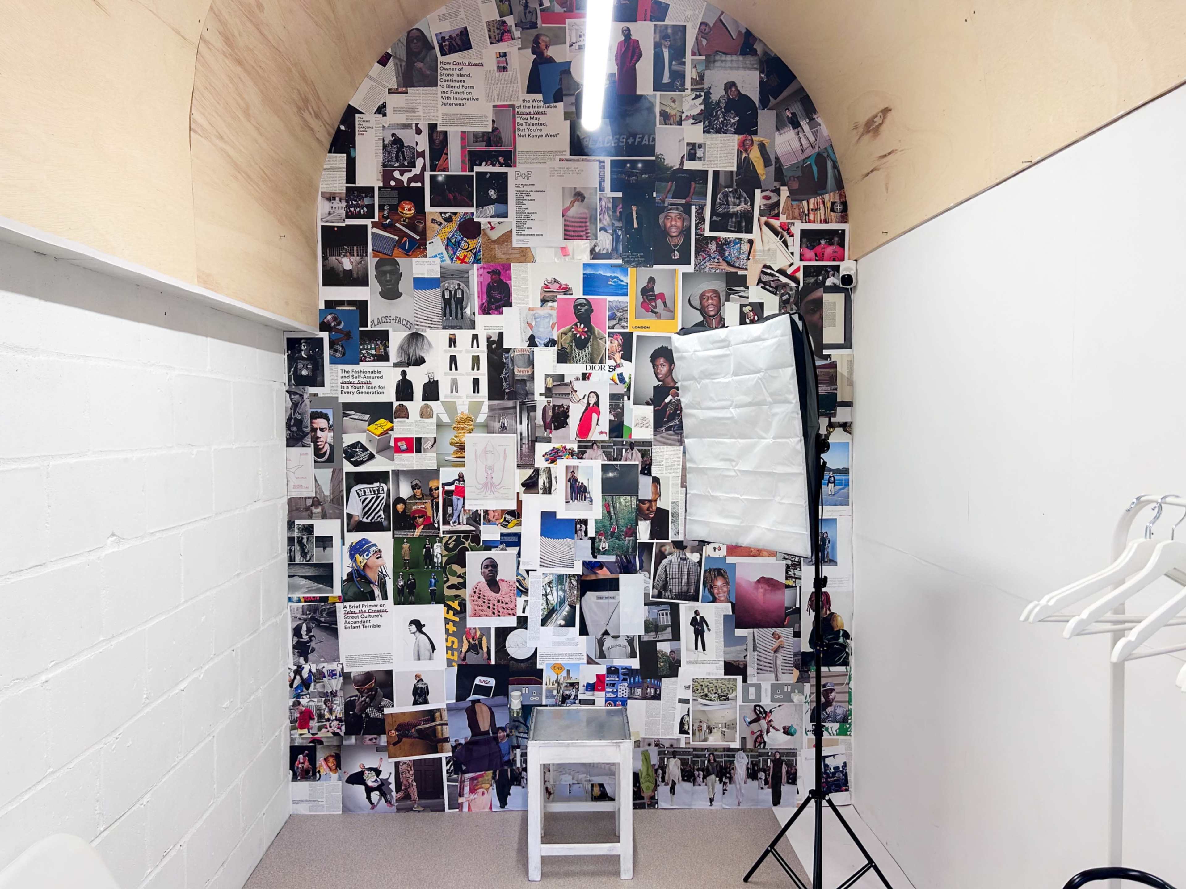 The image shows a small room with an arched wall covered in various photographs and art pieces, alongside a white backdrop and a light setup on a stand.