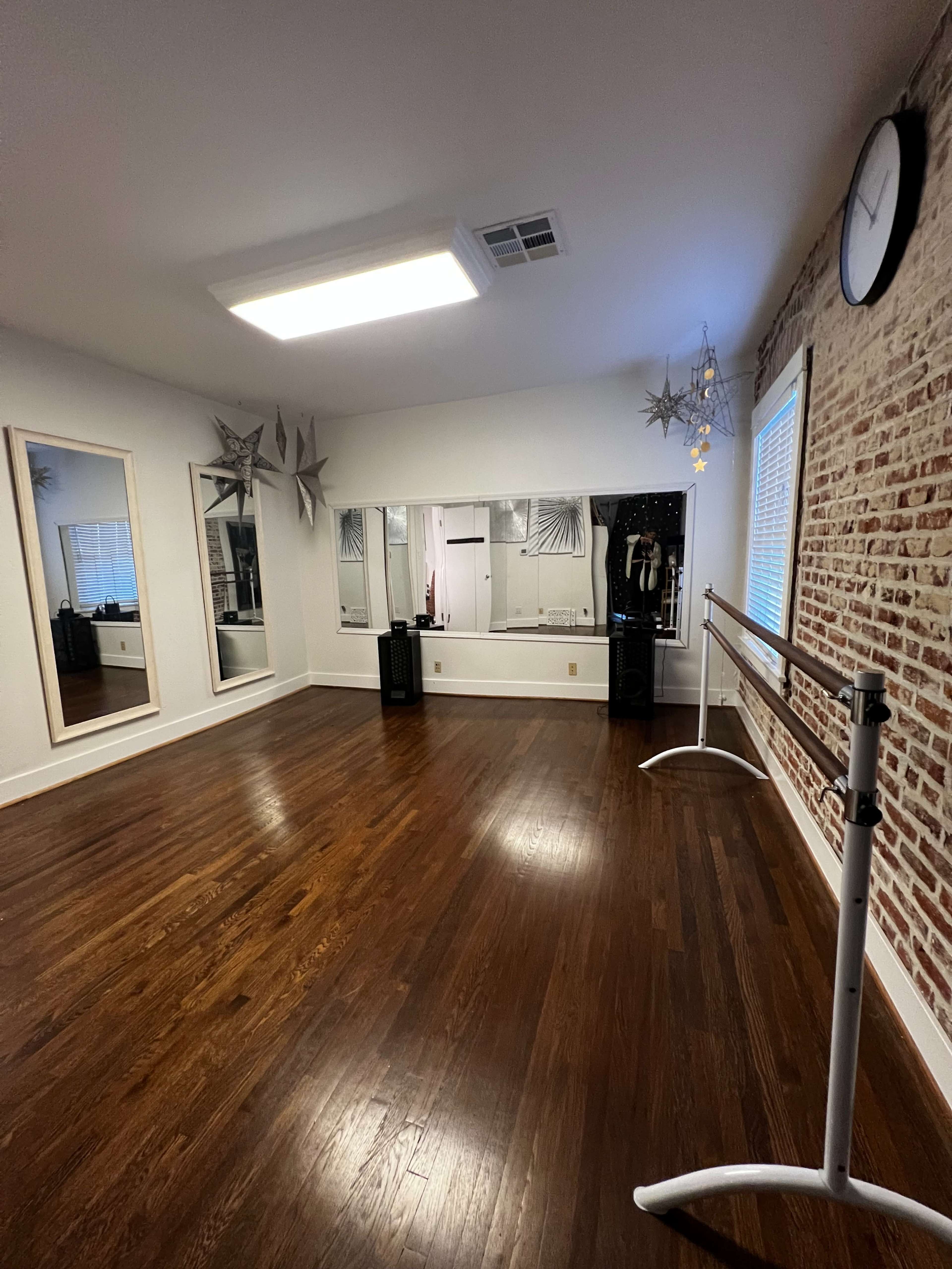 A dance studio with wooden flooring, decorative mirrors, a ballet barre, and a large illuminated ceiling fixture.