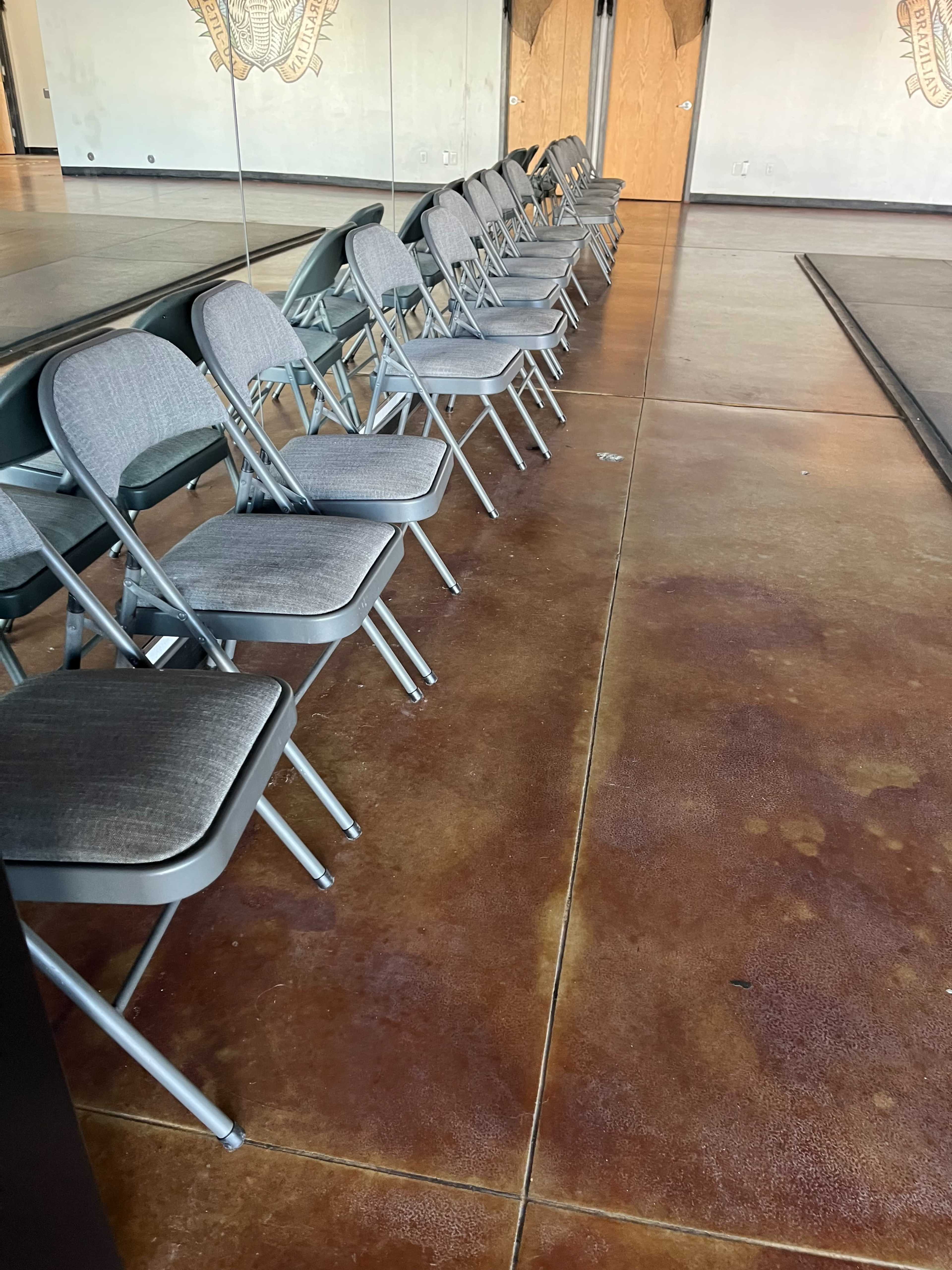 A row of gray folding chairs is positioned along a mirrored wall in a studio space with a polished concrete floor.