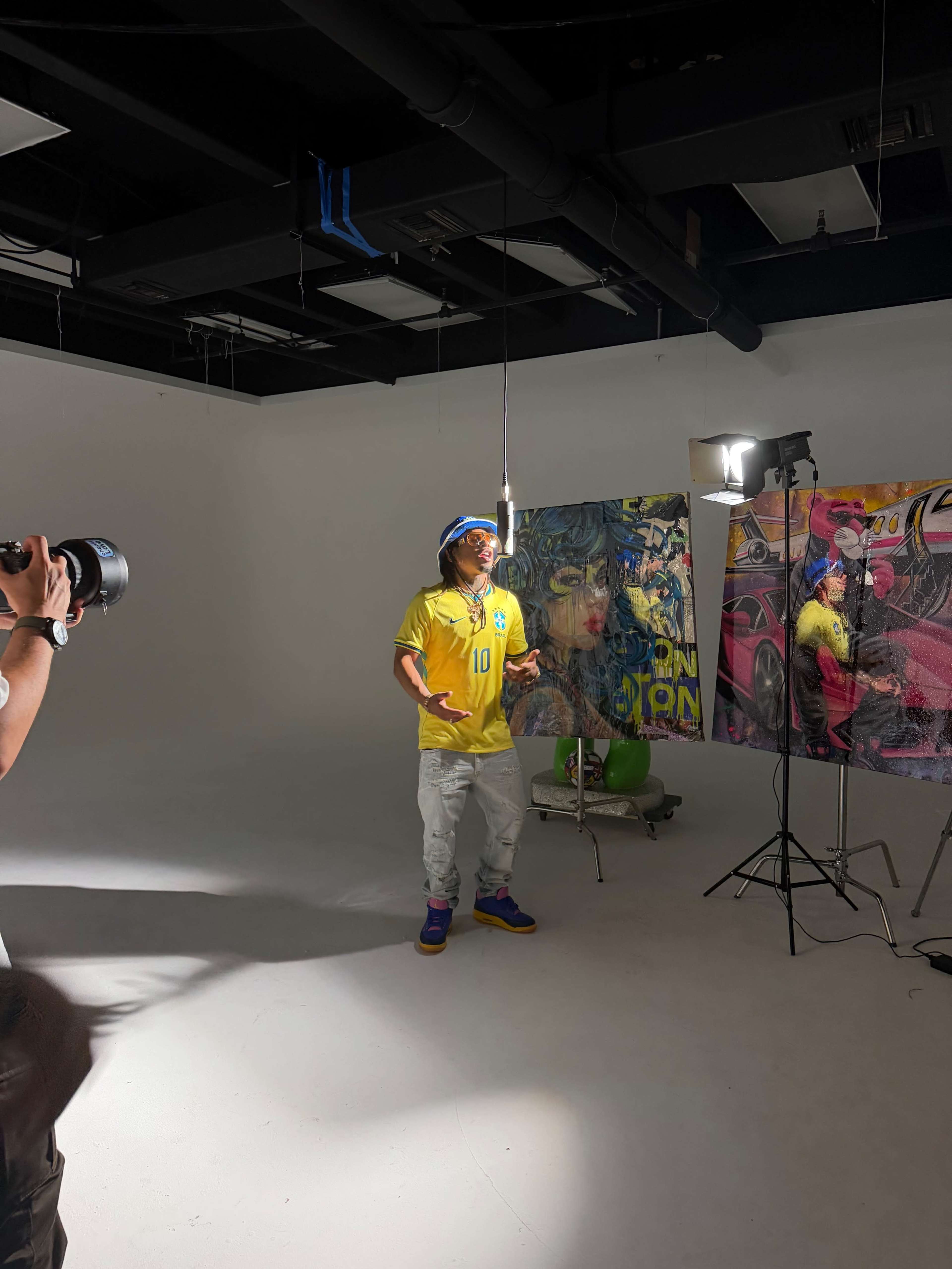 A person in a yellow jersey stands in a brightly lit studio with large, colorful artworks displayed nearby, while a photographer captures the scene.