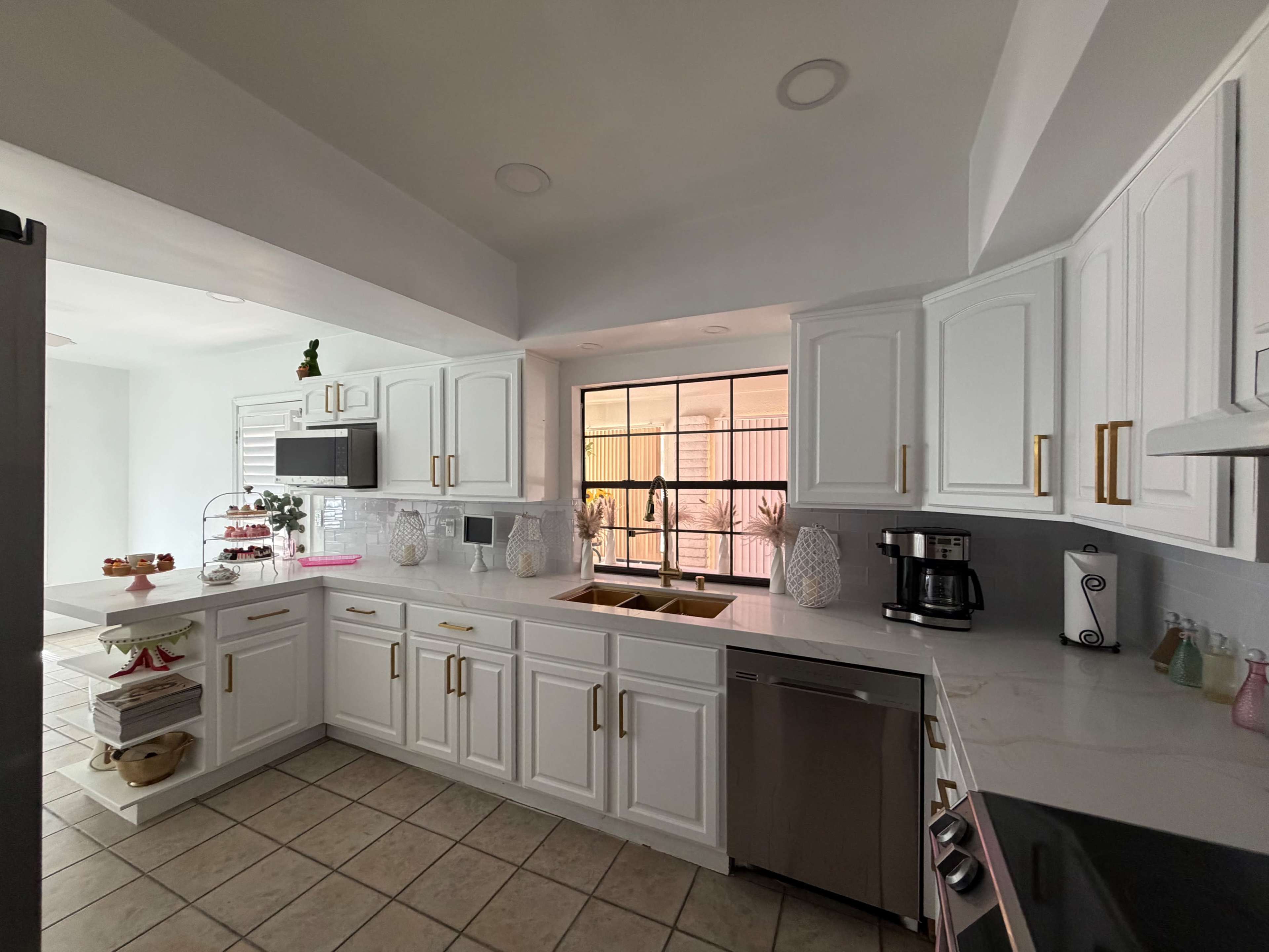 The image shows a modern kitchen with white cabinets, a stainless steel dishwasher, and a view of a window that lets in natural light.