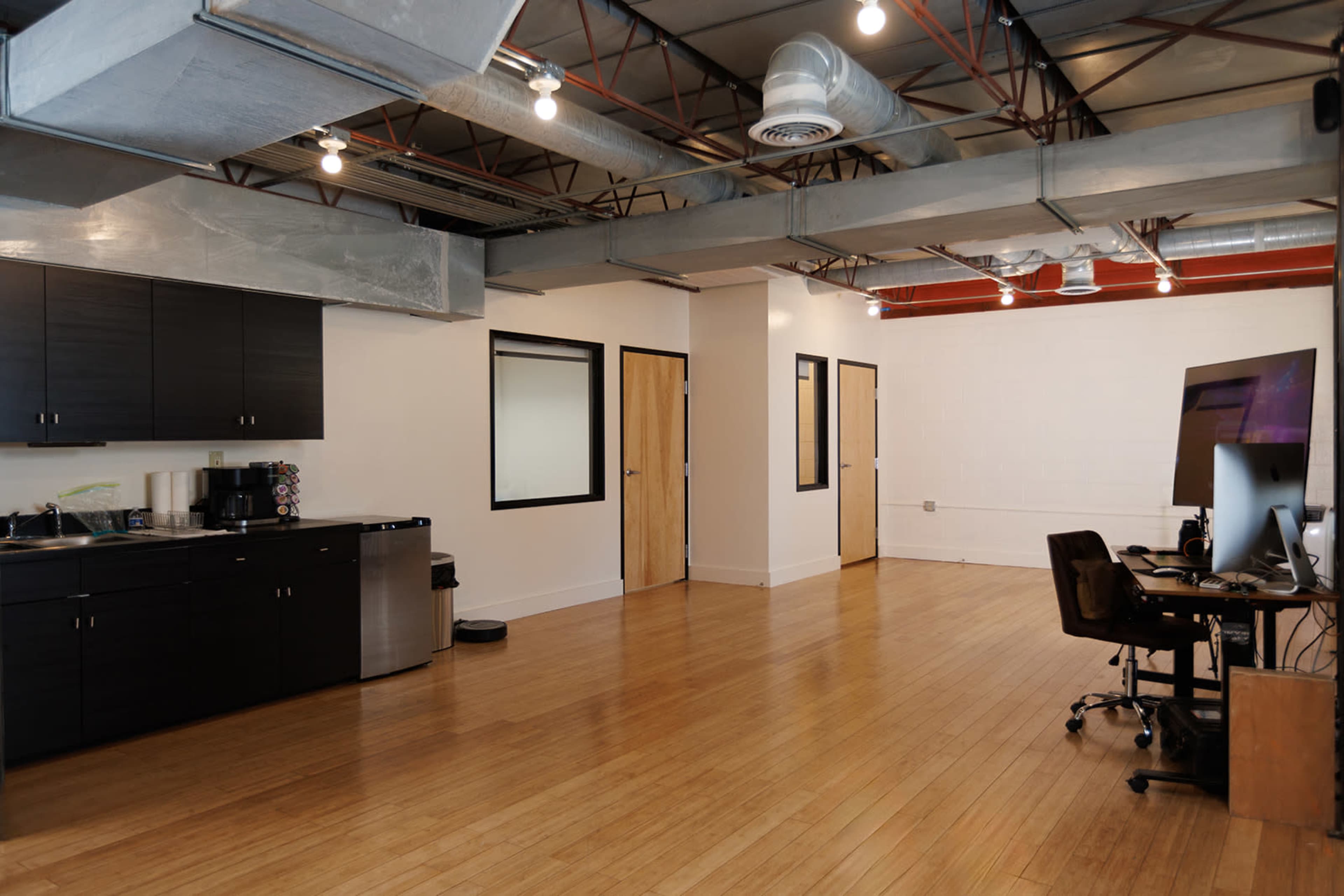 The image shows a modern, open office space with wooden flooring, some kitchen amenities, and a computer workstation against a wall.