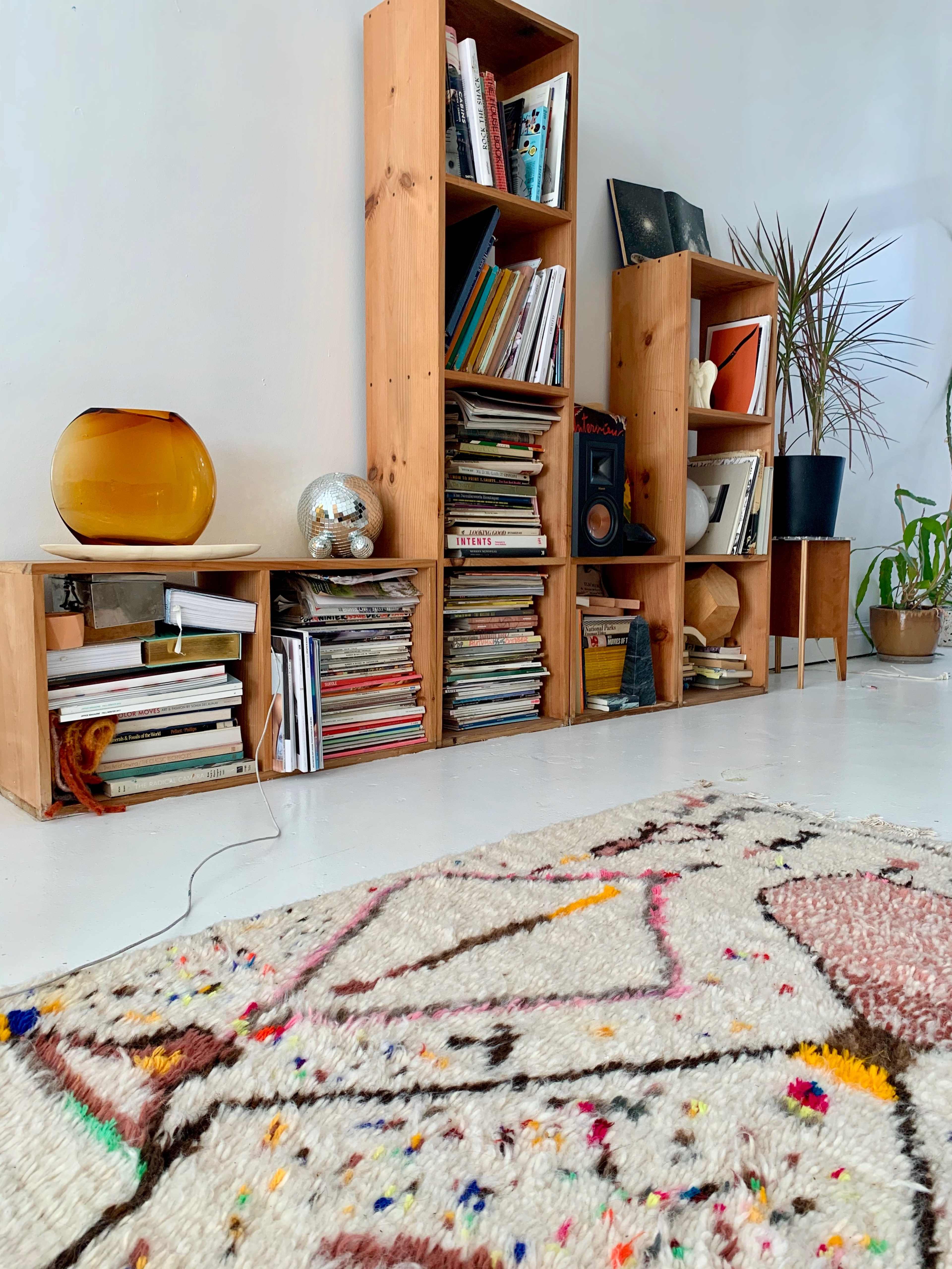 The image shows a cozy living space featuring wooden bookshelves filled with books, a decorative orange glass lamp, and a colorful patterned rug on the floor.