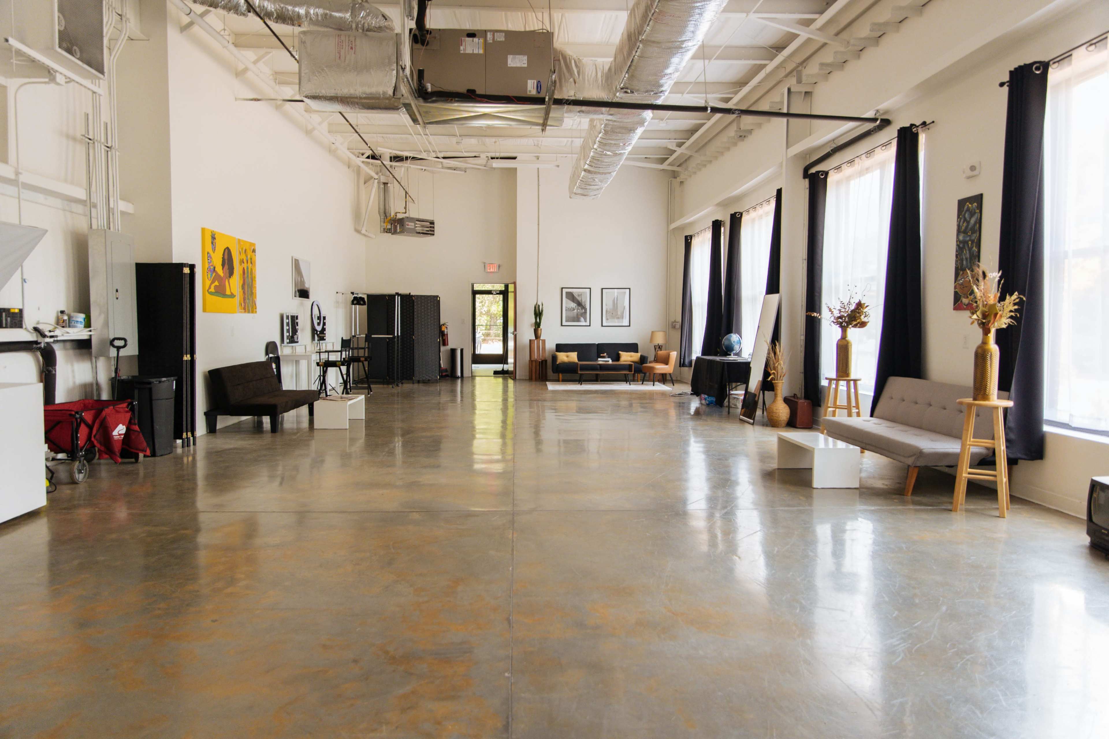 A spacious, well-lit interior of a room with a polished concrete floor, featuring seating areas and large windows with dark curtains.