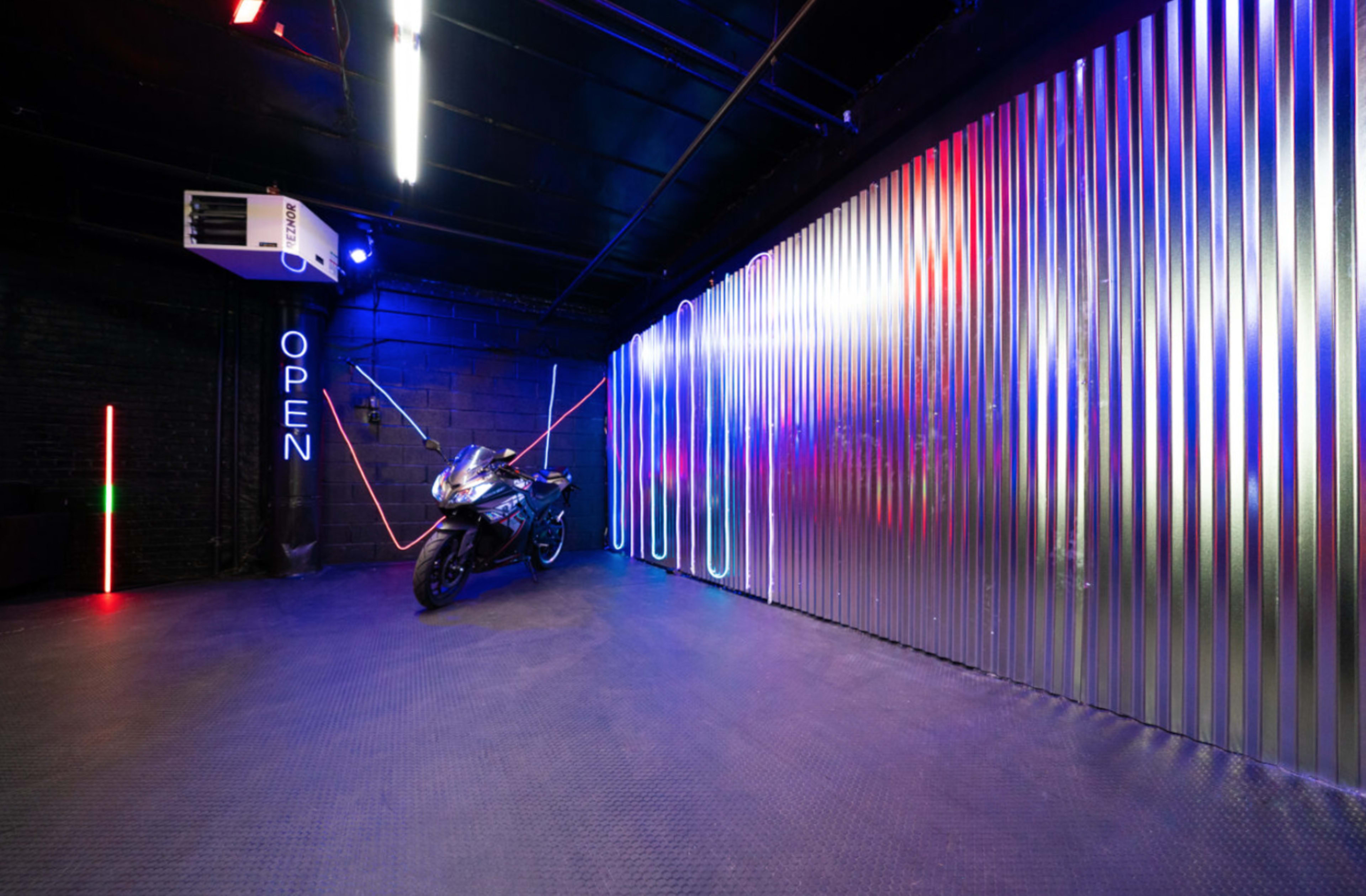 A motorcycle is positioned against a corrugated metal wall illuminated by colorful LED lights in a dimly lit room.