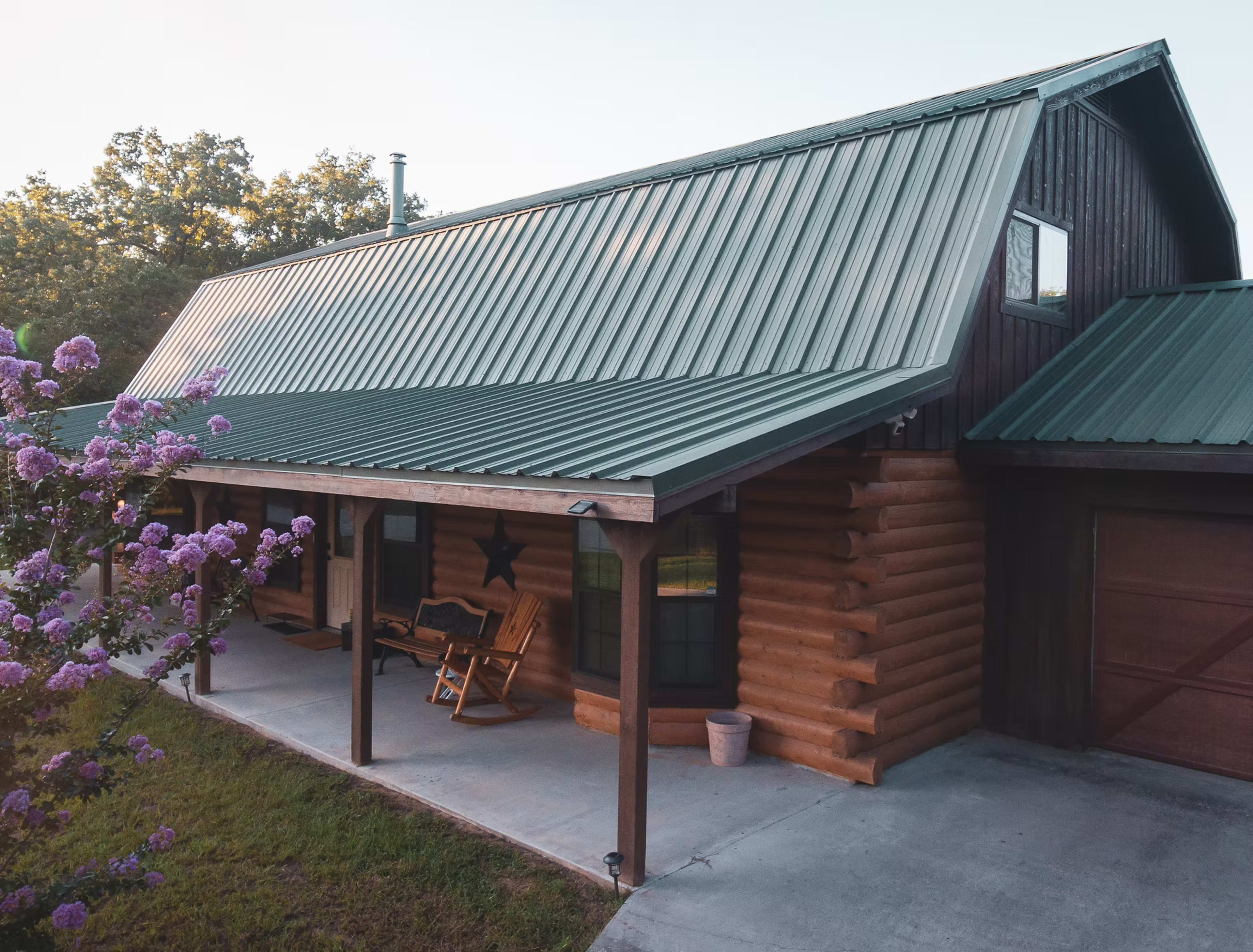 The image shows a log cabin with a green metal roof and a porch, surrounded by grass and flowering plants.