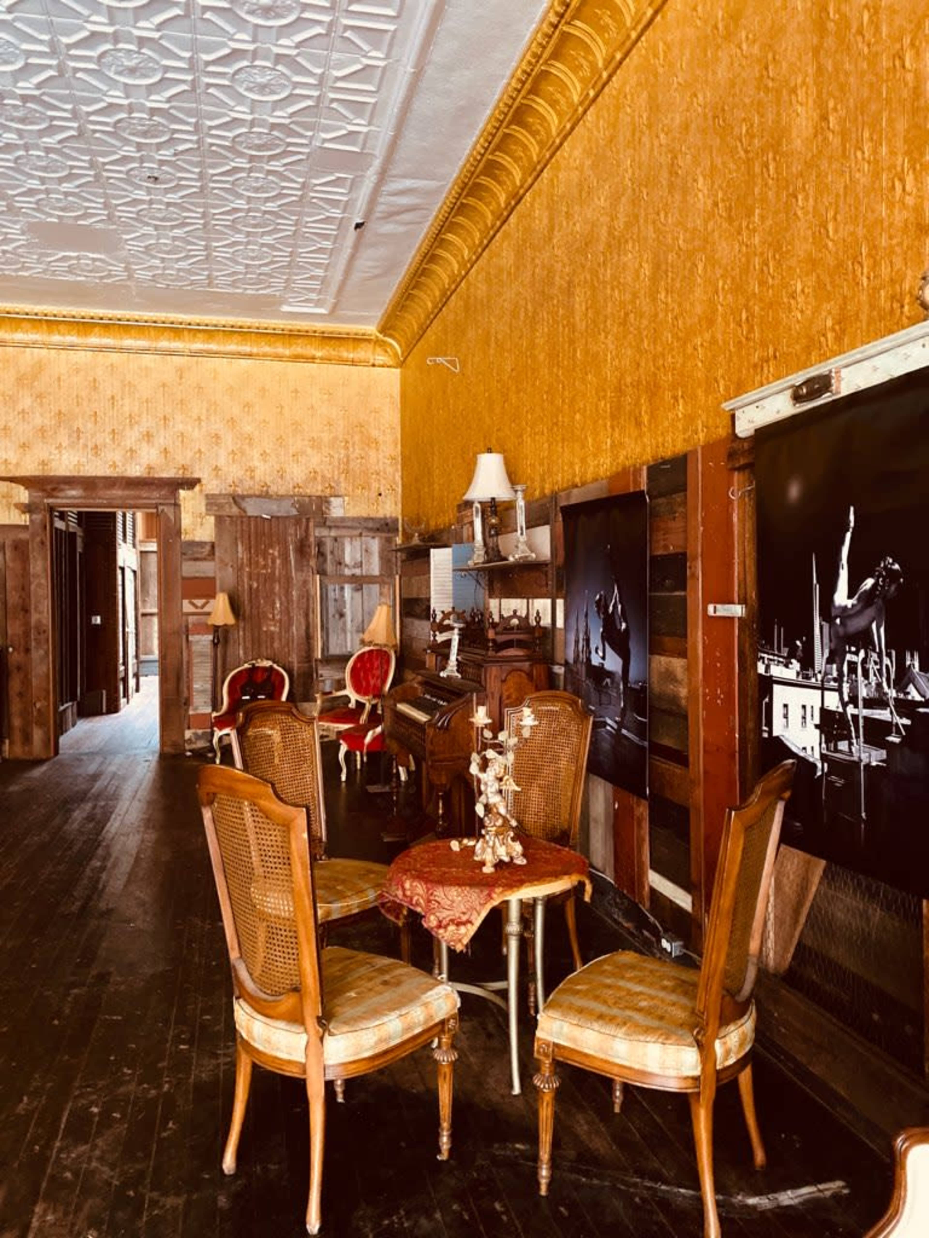 A vintage room with wooden flooring, adorned with gold wallpaper and antique furniture, including a table with two chairs and a decorative centerpiece.