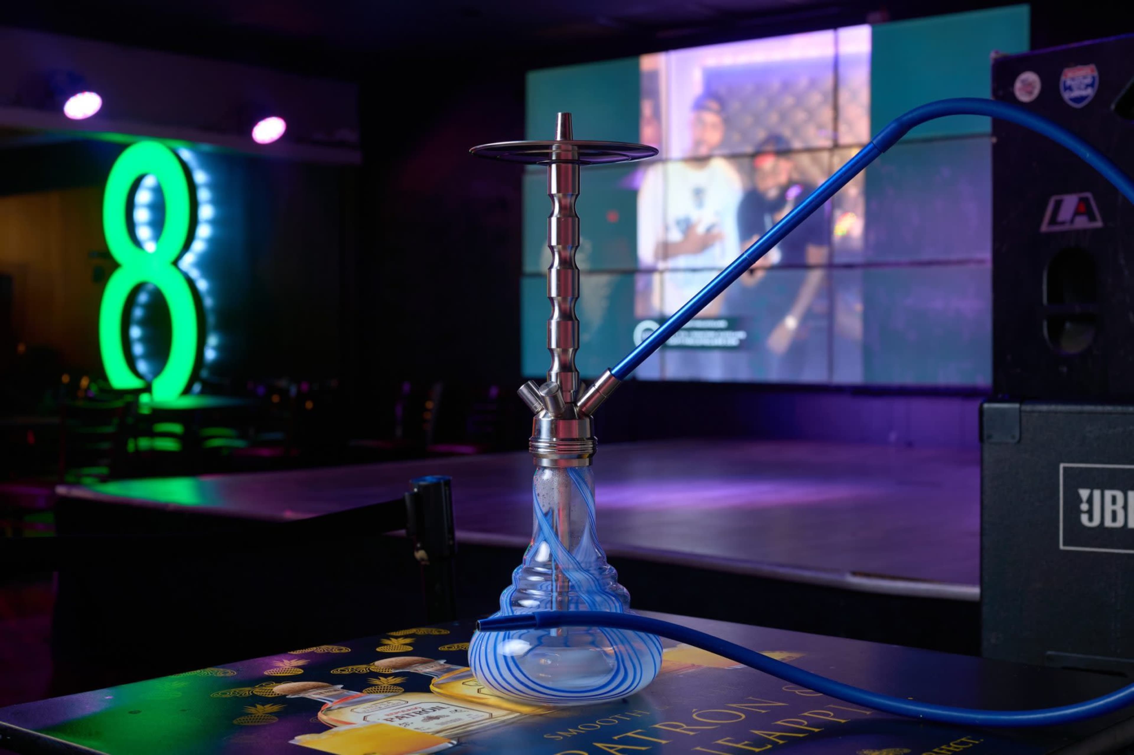 A hookah with a blue hose is placed on a table in a dimly lit venue featuring a large screen in the background displaying a video.