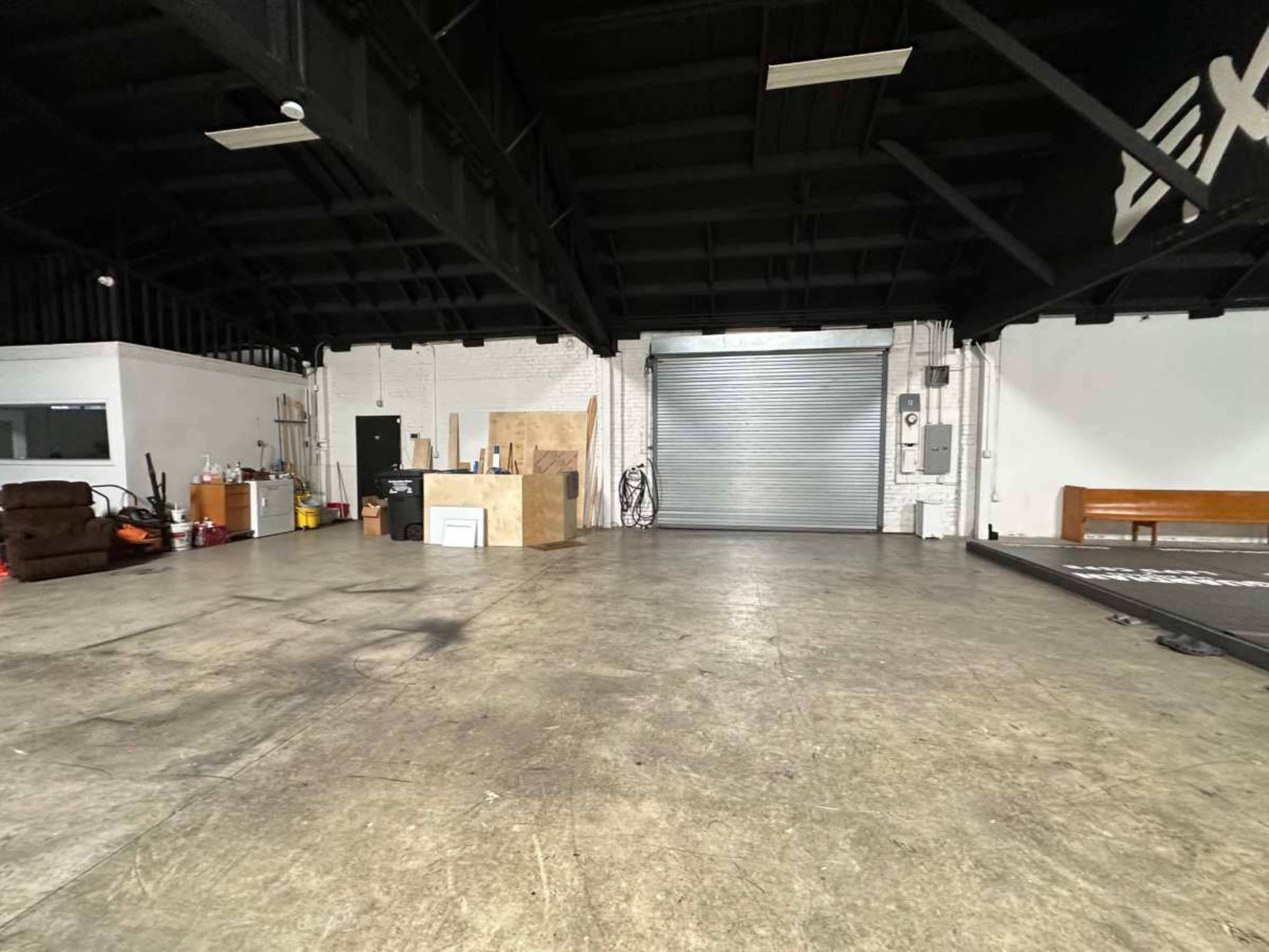 The image shows an empty industrial space with a concrete floor, a closed metal roll-up door, and various items arranged against the walls.
