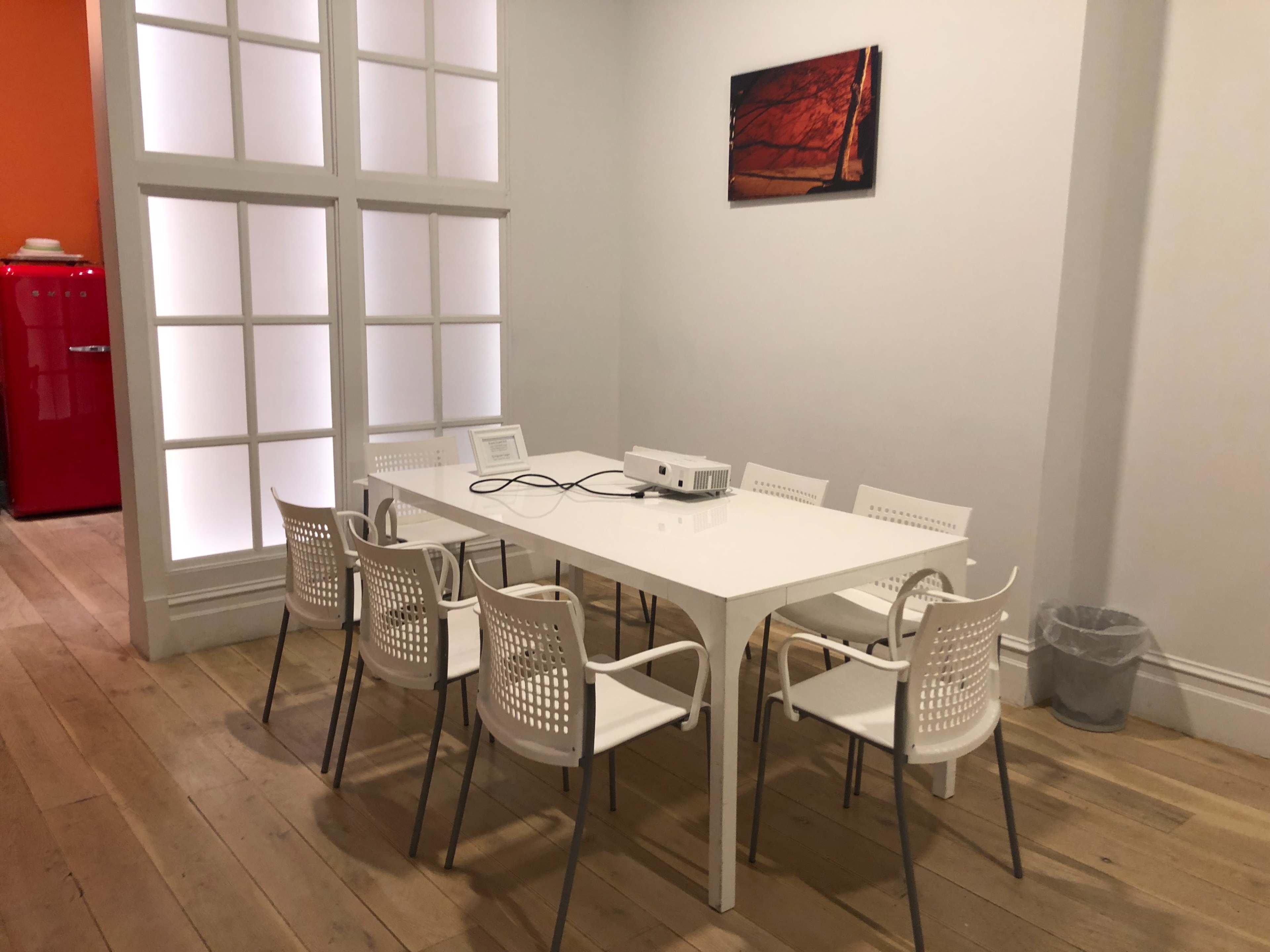 A conference room features a white table surrounded by six chairs, a projector on the table, and a red fridge visible in the background.