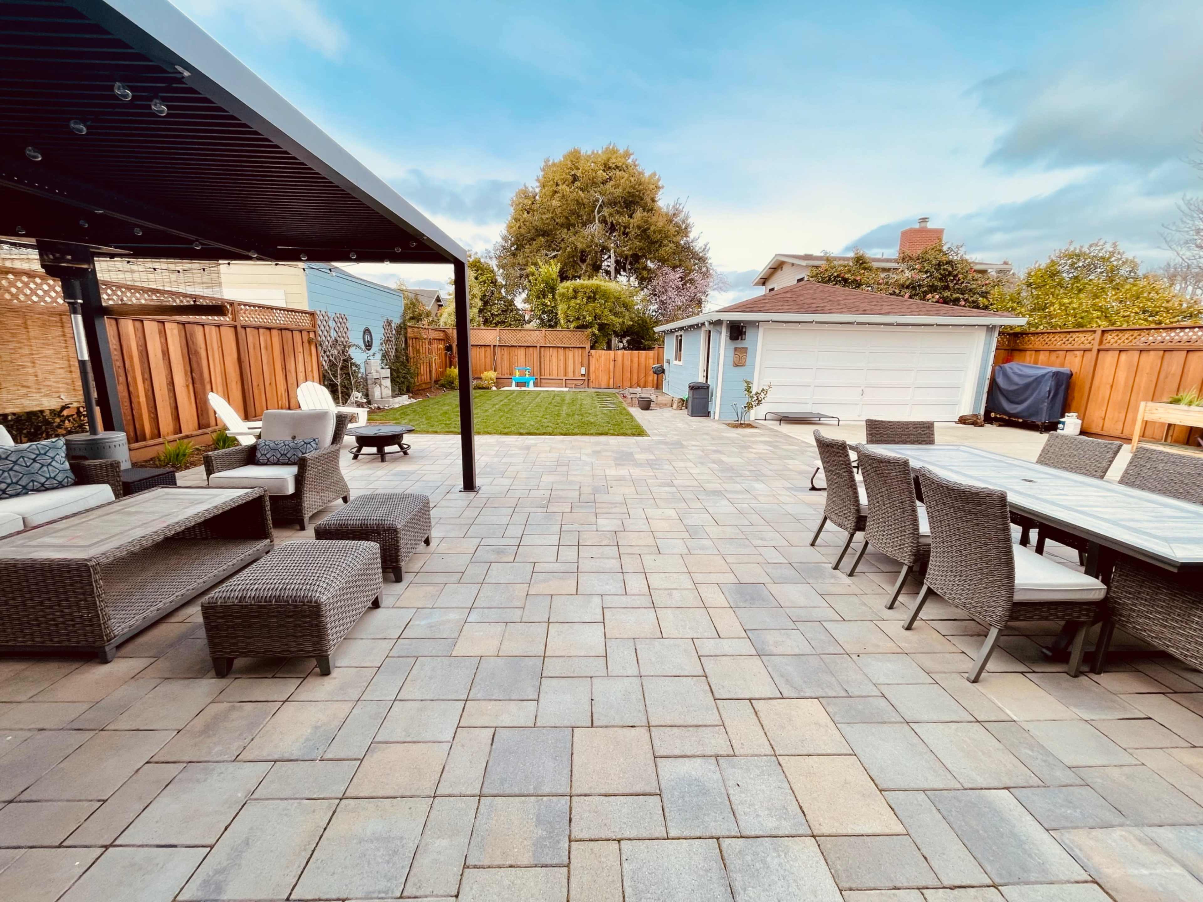 A spacious backyard patio area with a combination of seating arrangements and a grassy lawn, surrounded by wooden fencing and a garage in the background.