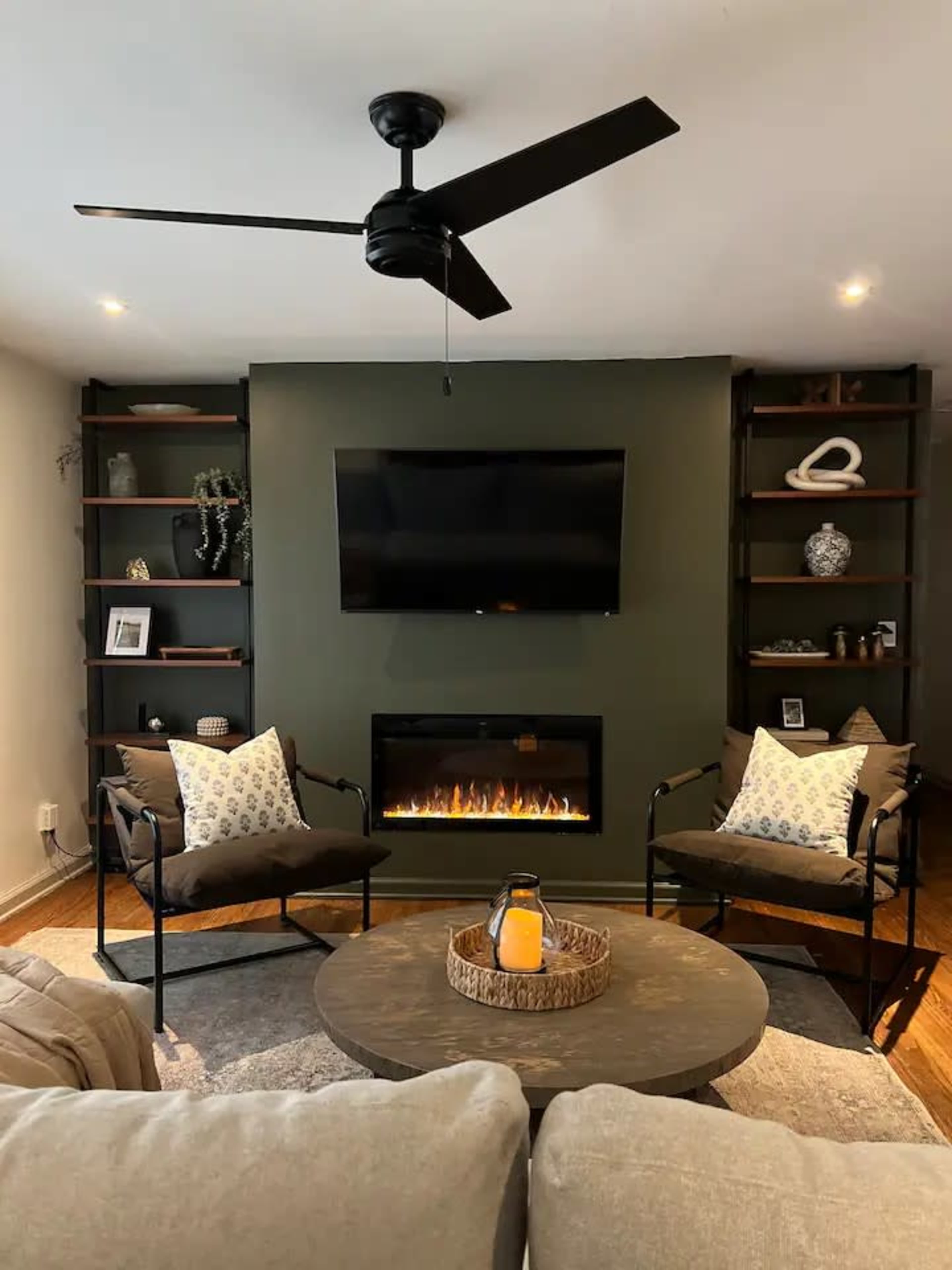 The image shows a cozy living room featuring a modern electric fireplace beneath a flat-screen TV, flanked by dark wooden shelves filled with decorative items, and two chairs with patterned pillows facing a circular coffee table.