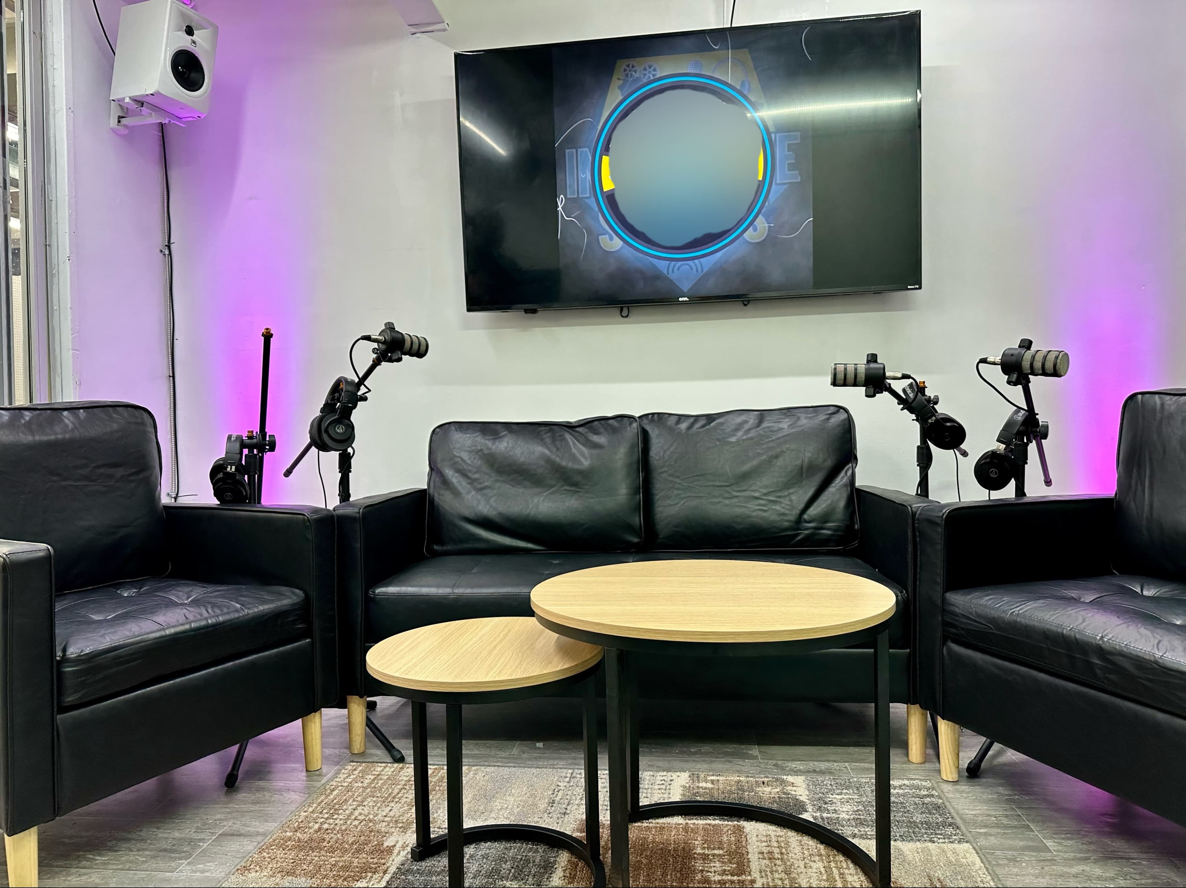 A seating area with two black couches facing a wall-mounted screen, flanked by microphones on stands.