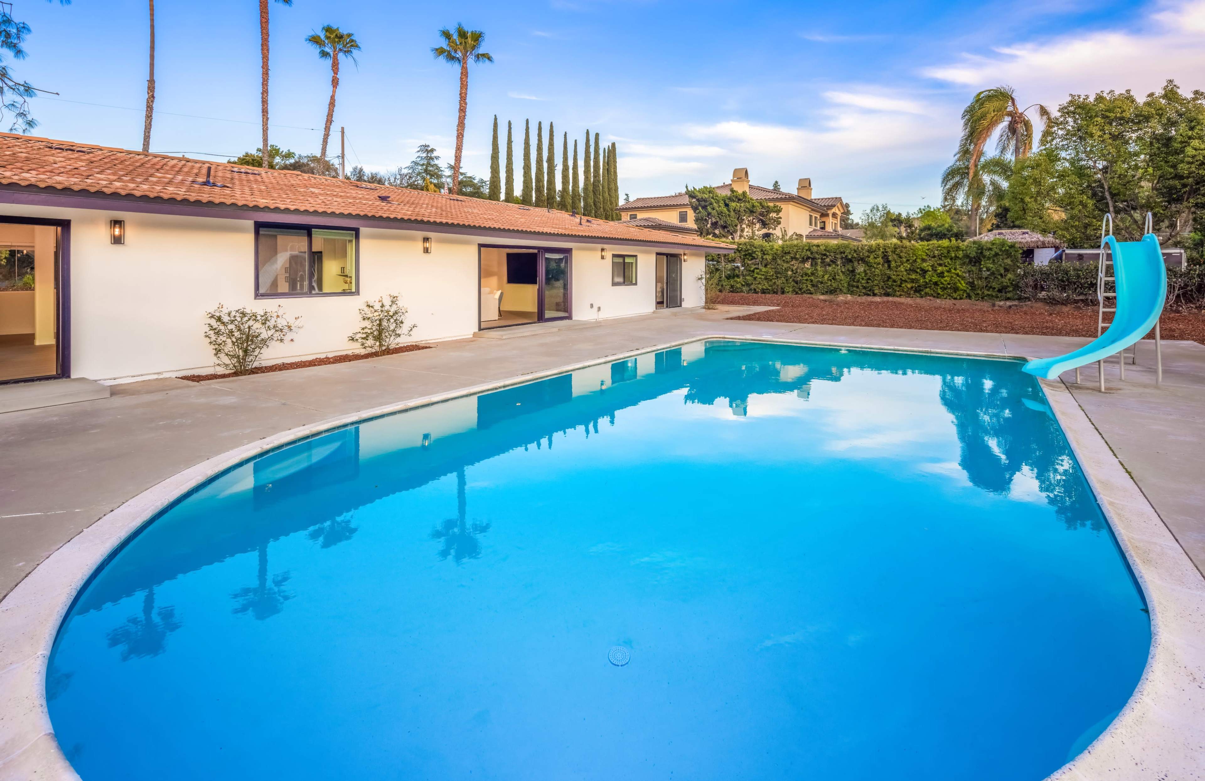 A residential backyard featuring a swimming pool with a slide and a single-story house surrounded by palm trees and manicured landscaping.