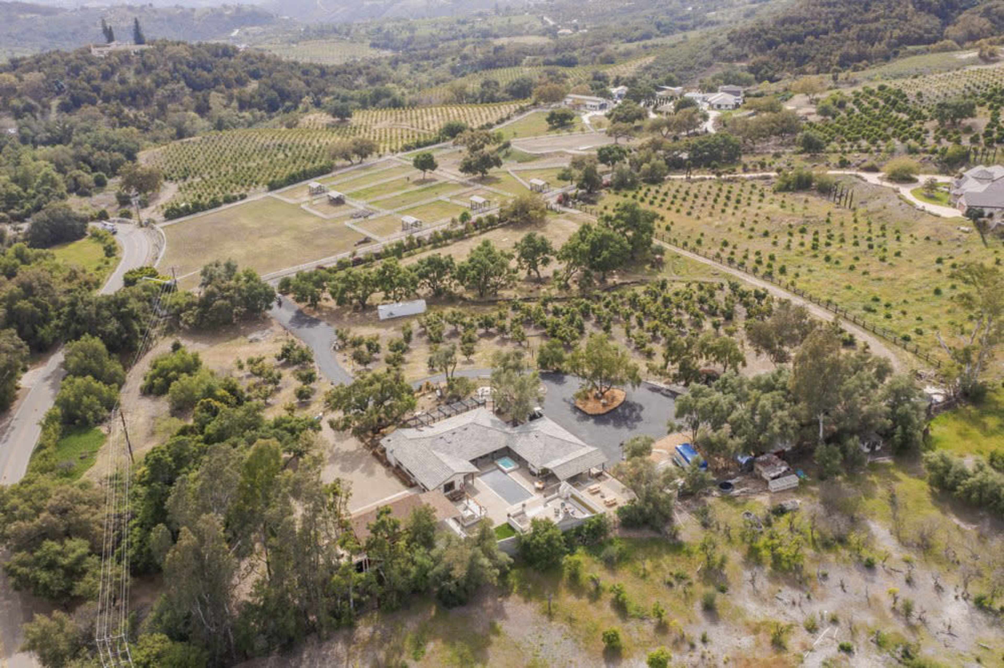 The image shows an aerial view of a large property featuring a house, a swimming pool, and expansive greenery, including orchards and vineyards, situated in a rural landscape.