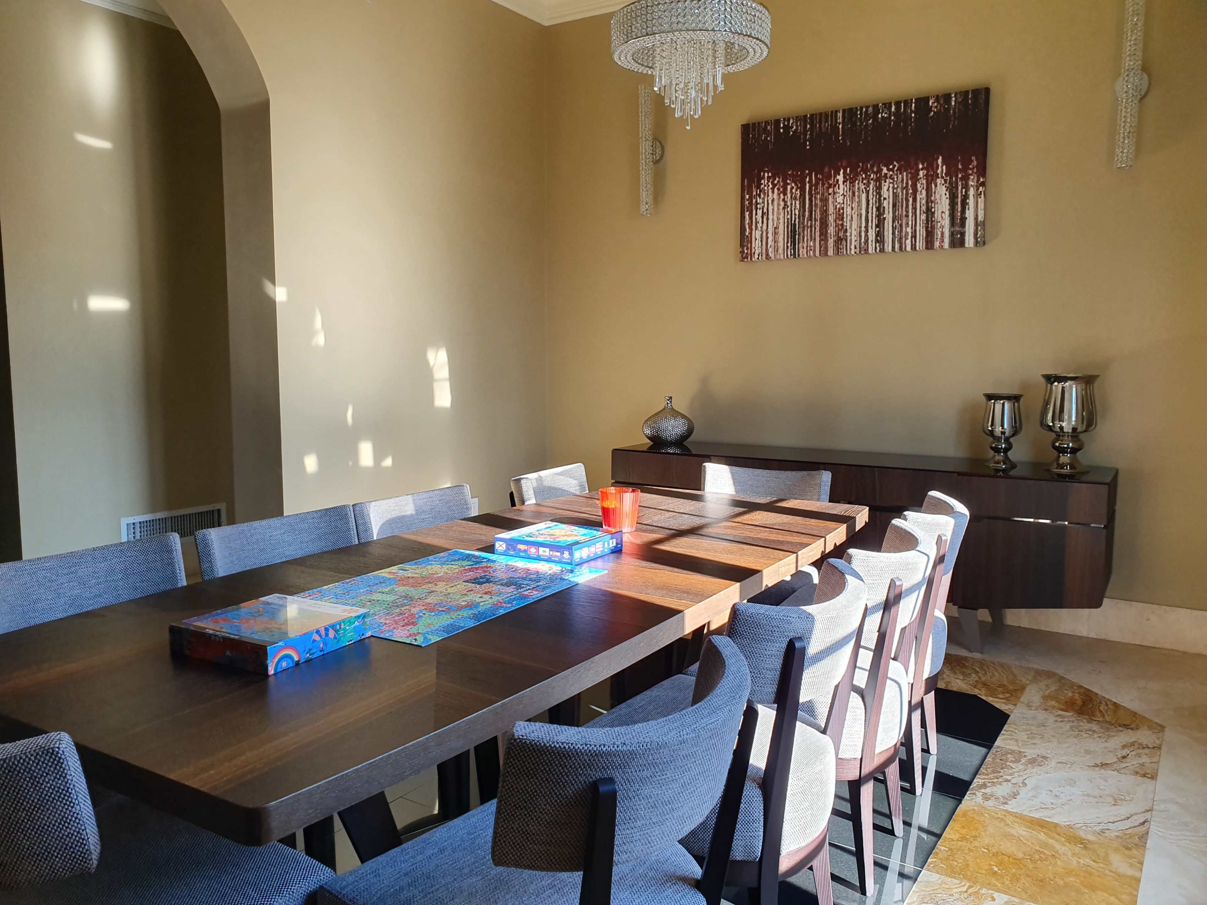 A spacious dining room features a large wooden table surrounded by chairs, with a puzzle and a candle on the table, and a sideboard displaying decorative items.