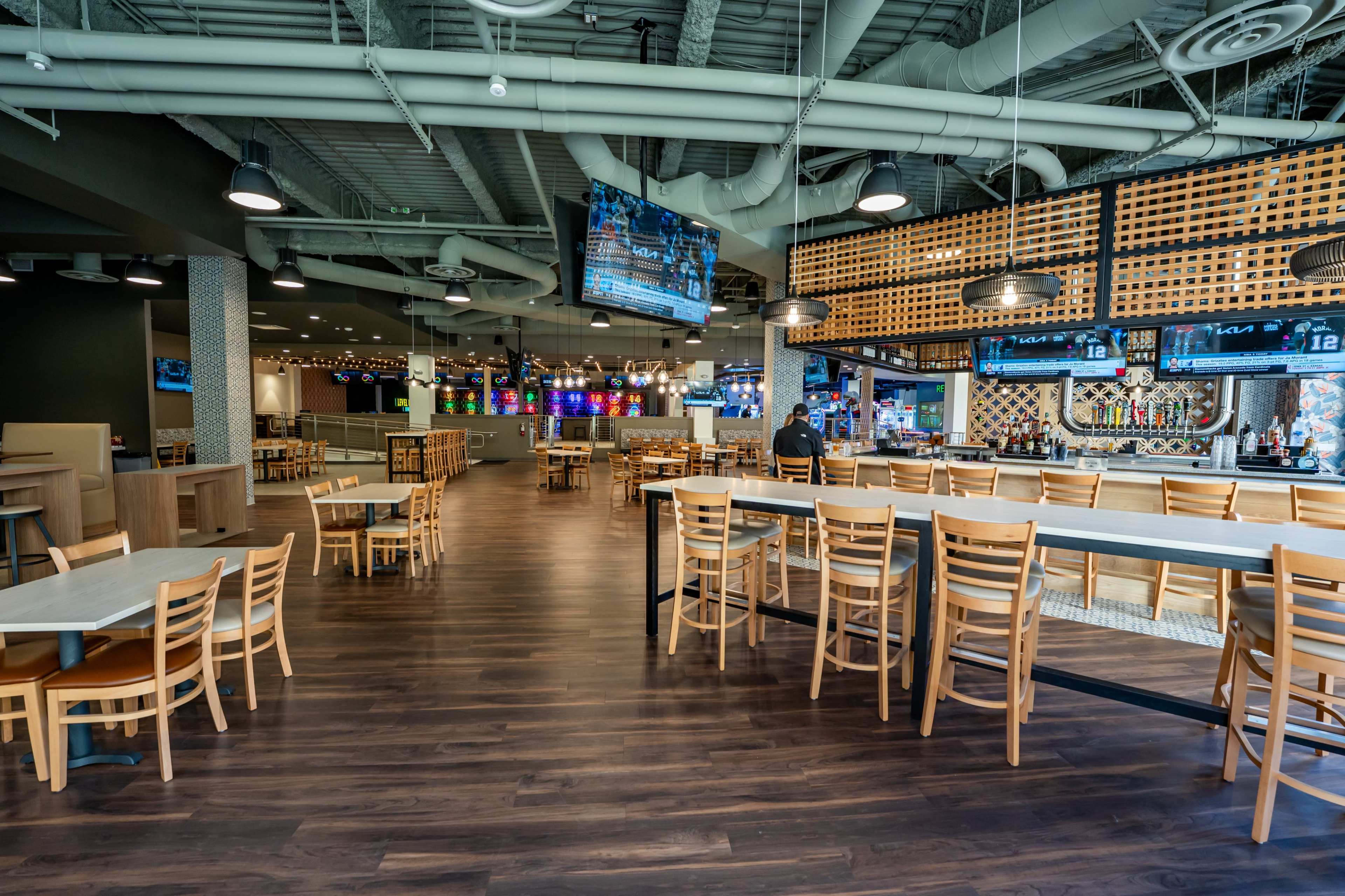 The image shows a spacious, modern dining area with numerous tables and chairs, a bar with multiple screens, and an open layout.