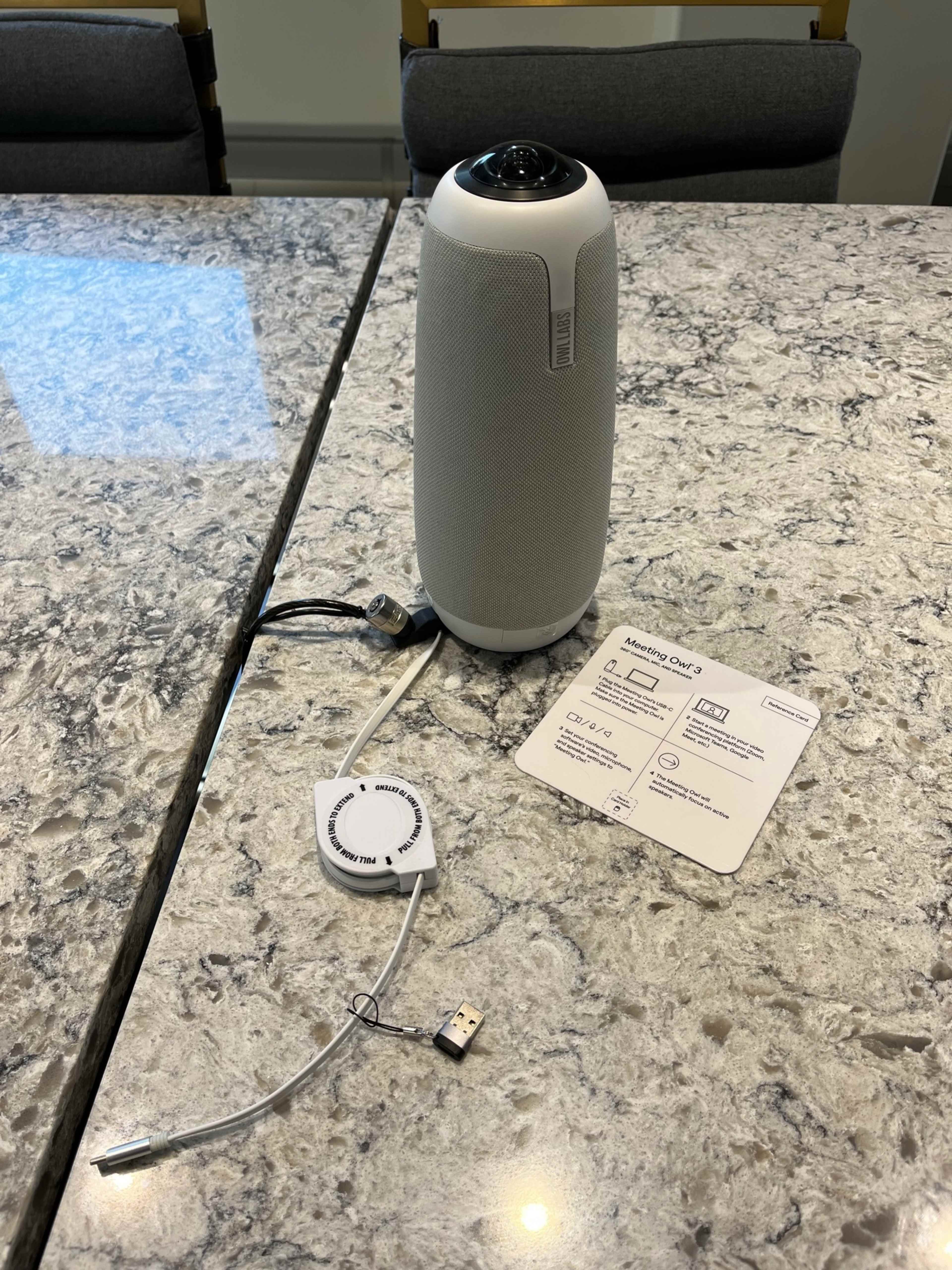 A smart speaker is placed on a marble countertop next to a user guide and charging accessories.