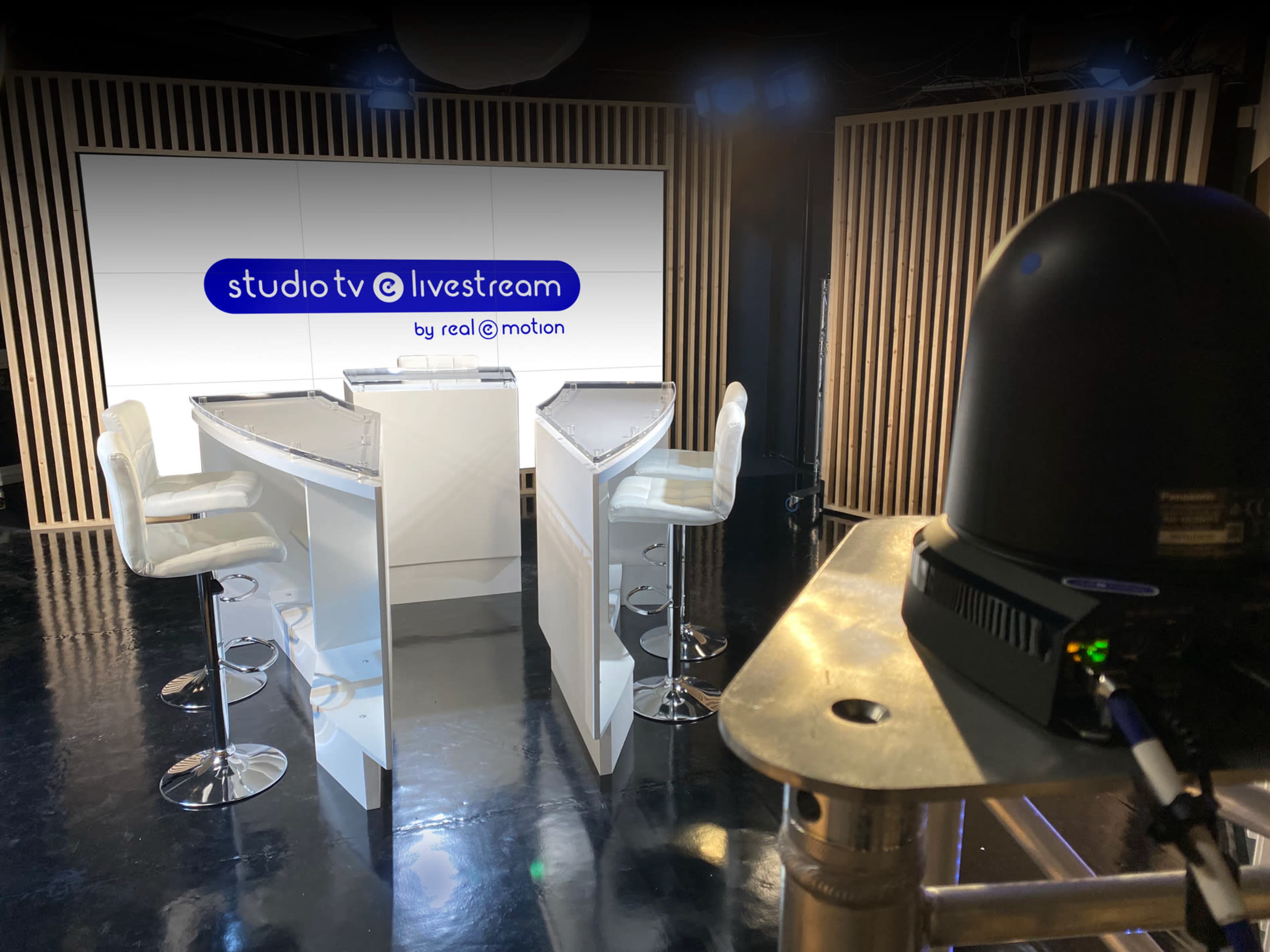 A modern television studio setup with two sleek, white podiums and four bar stools against a backdrop featuring a lit screen displaying the text "studio tv @ livestream by real @ emotion."